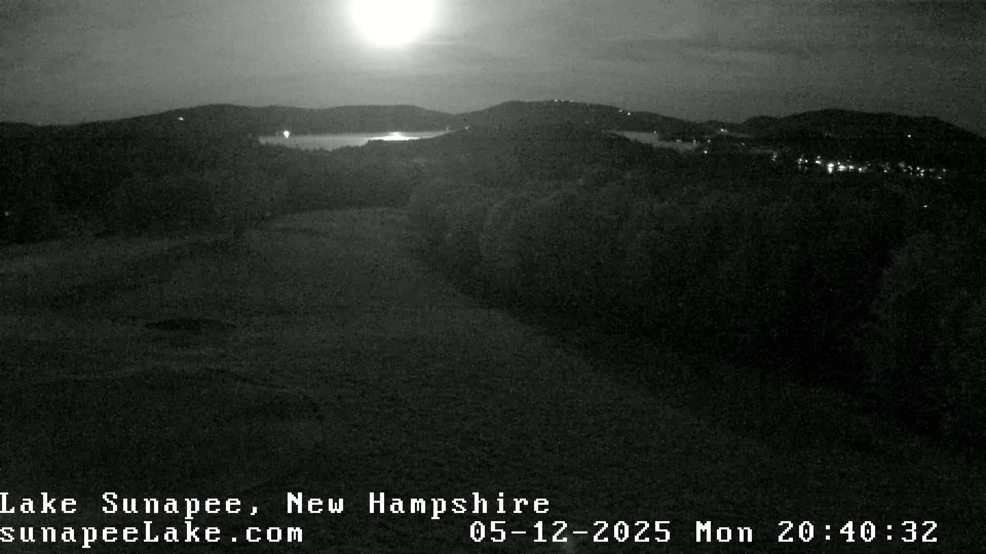 A nighttime view of a lake nestled between dark hills under a bright moon.