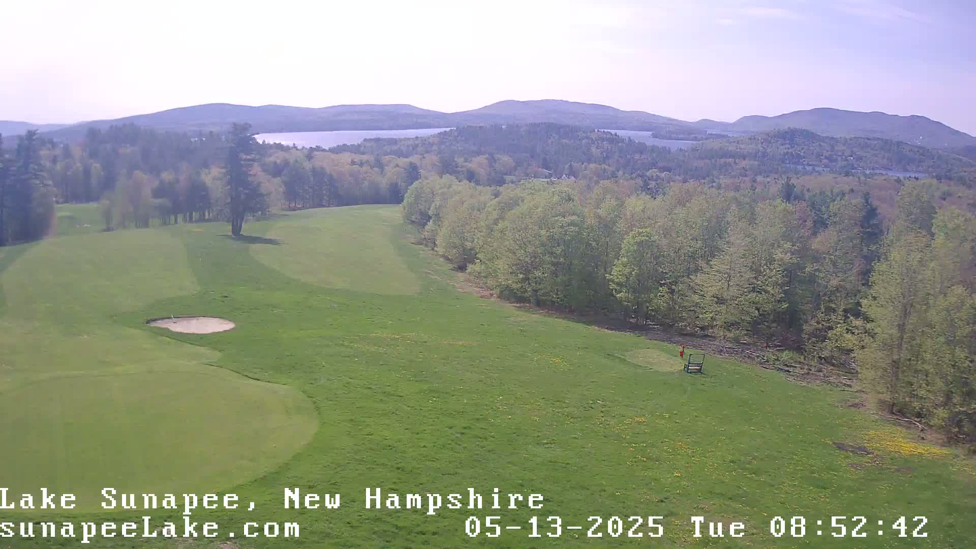 A golf course with a sand trap and a distant view of a lake and mountains under a bright, mostly clear sky.