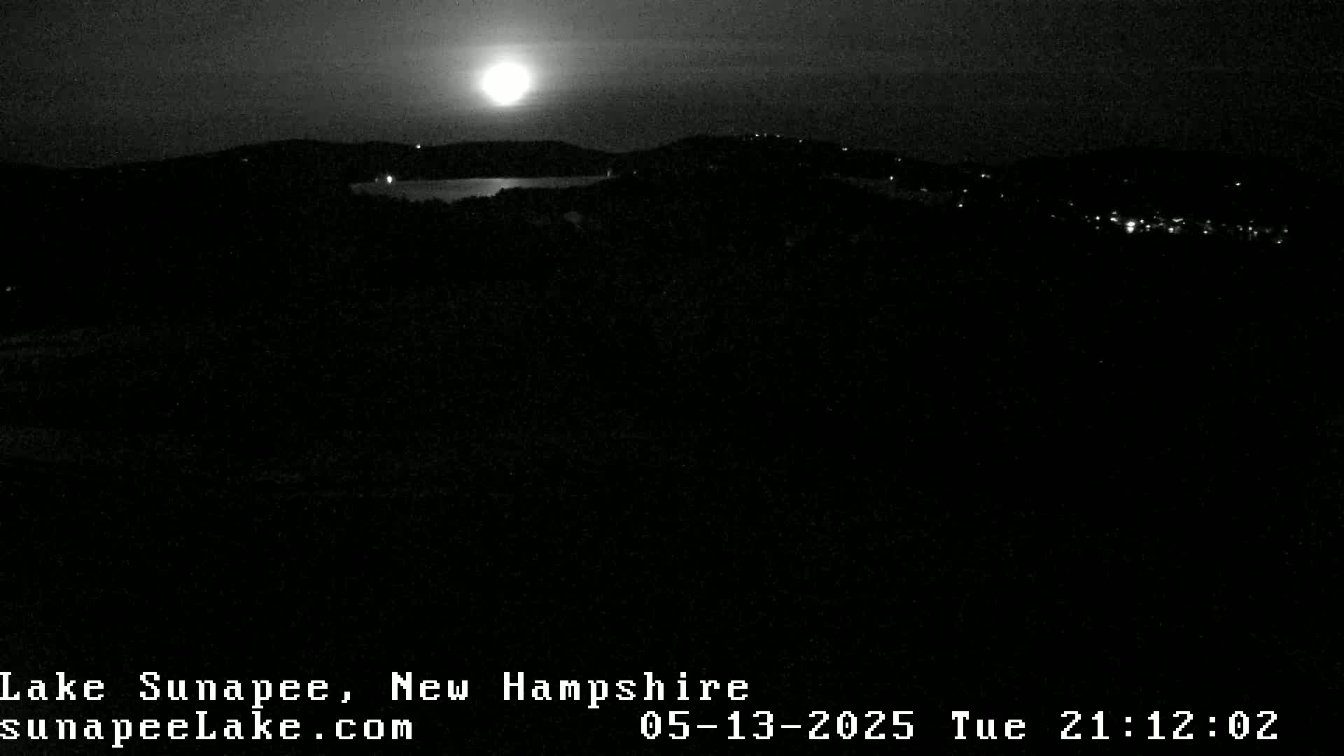 A bright moon illuminates a dark landscape featuring a body of water and distant lights at night.