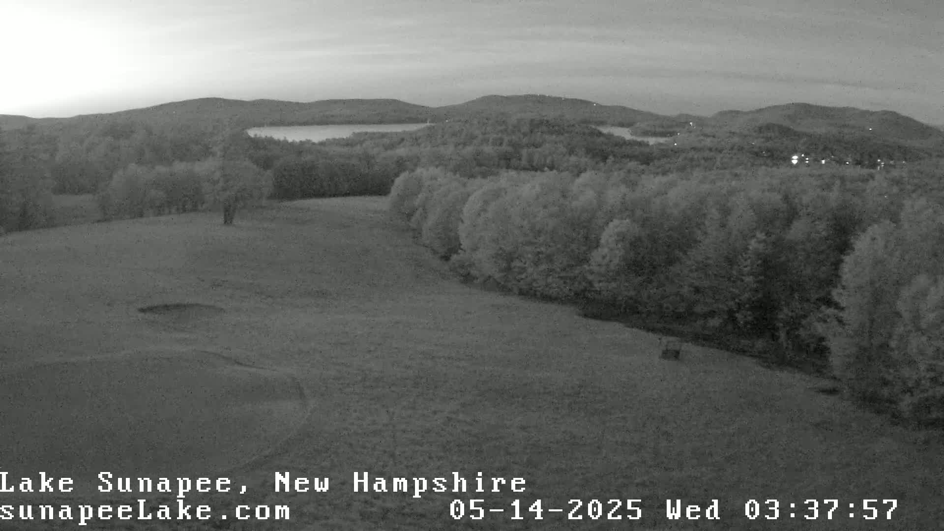 An elevated, grayscale view shows a grassy field sloping down to a wooded area, with a lake and distant hills visible in the background under a twilight sky.