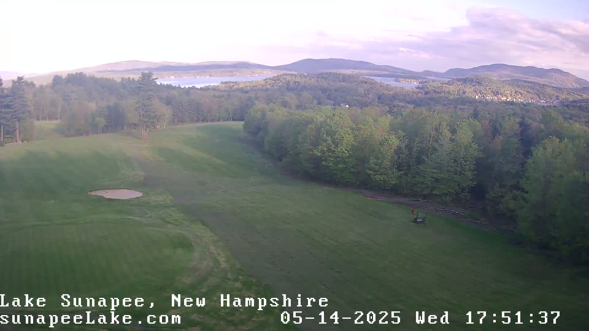 A high-angle view shows a mostly sunny, grassy golf course overlooking a lake and distant mountains.