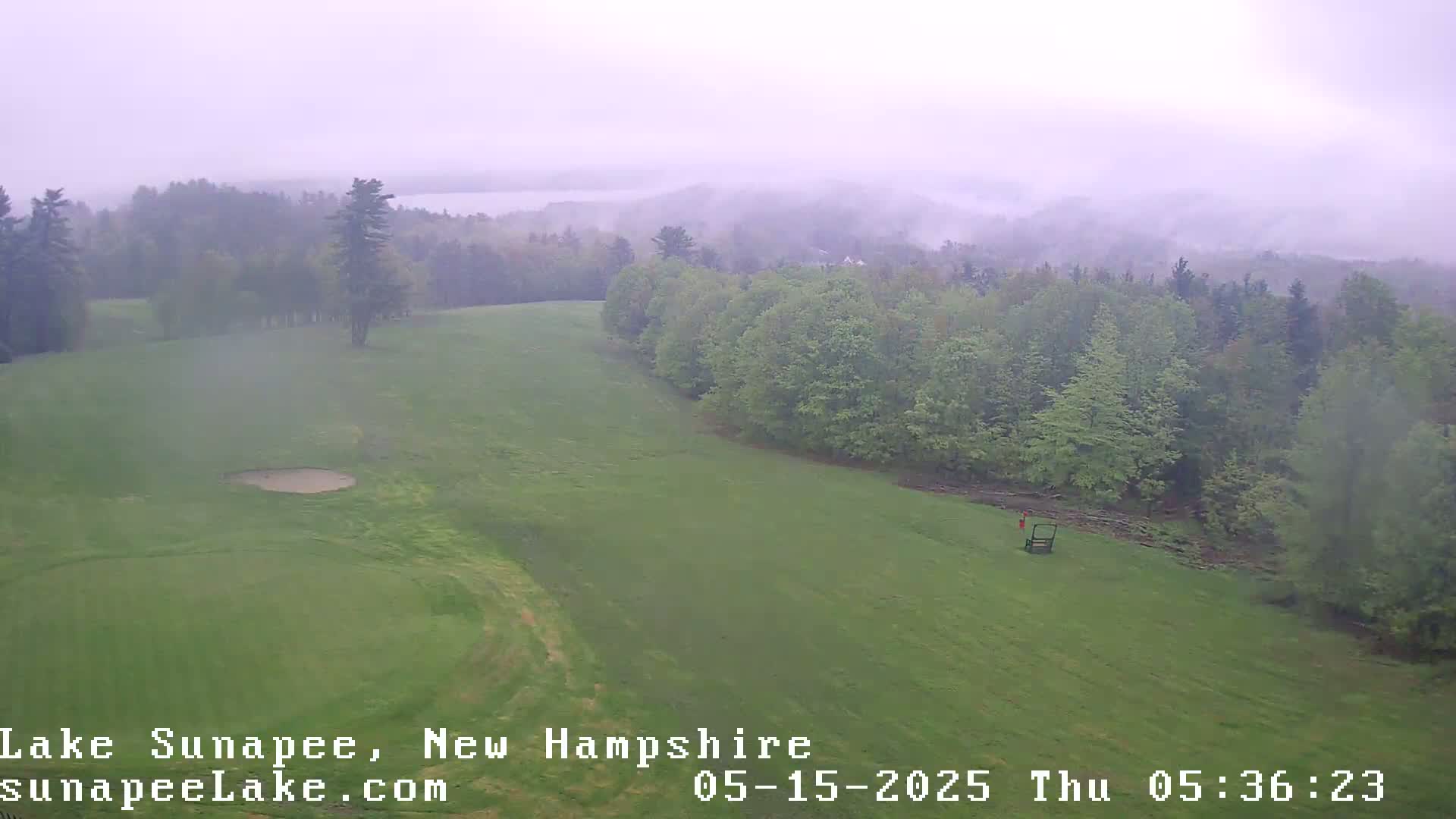 A misty, overcast view shows a grassy expanse of land, possibly a golf course, bordered by a line of green trees and a distant, hazy lake.