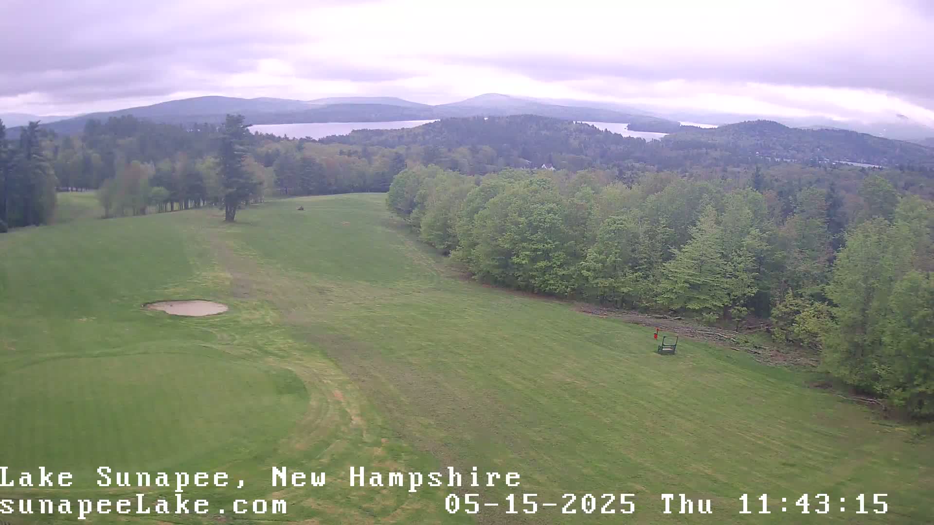 An aerial view shows a green golf course with a sand trap, bordered by a line of trees, overlooking a lake and distant mountains under an overcast sky.