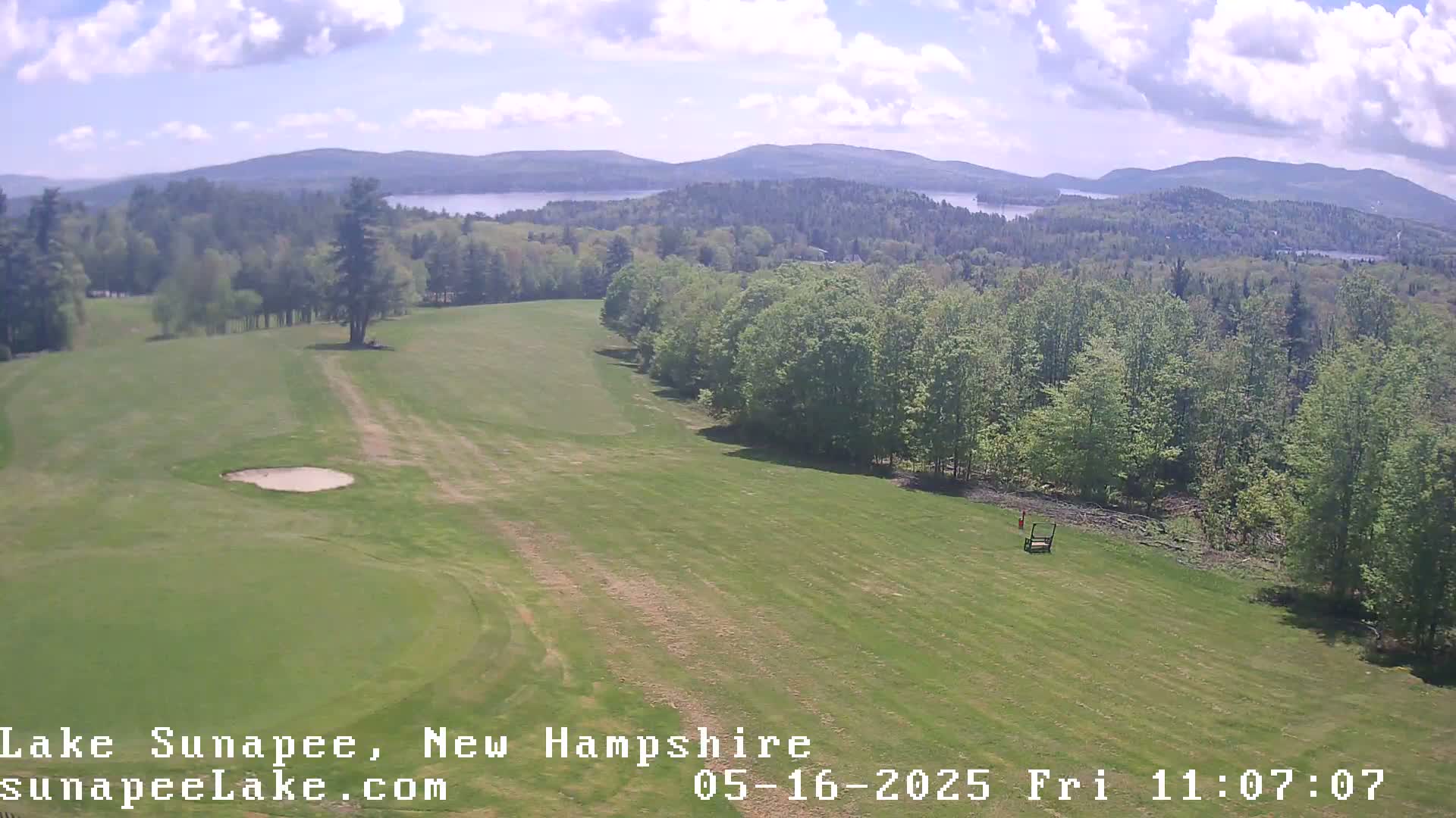 An aerial view shows a golf course fairway bordered by a forest, overlooking a lake and distant mountains under a partly cloudy sky.