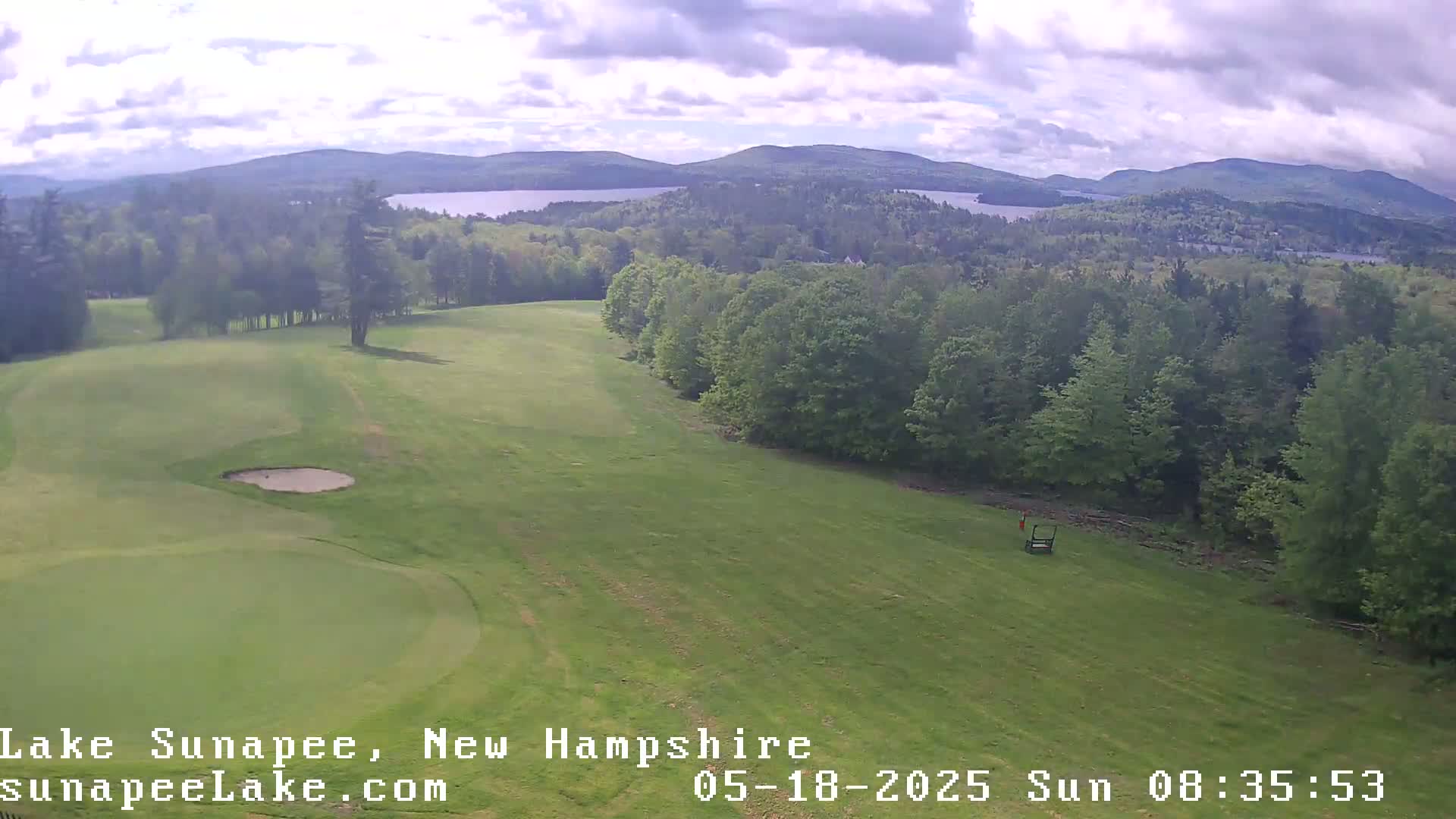 An aerial view shows a partly sunny day over a golf course with a lake and mountains in the distance.