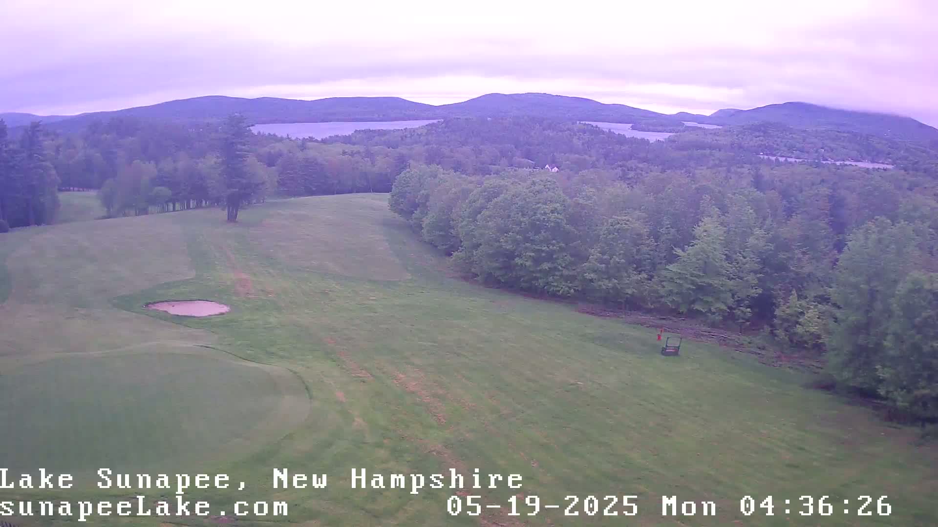 An aerial view of a golf course with a sand trap, overlooking a lake and mountains under a cloudy sky.