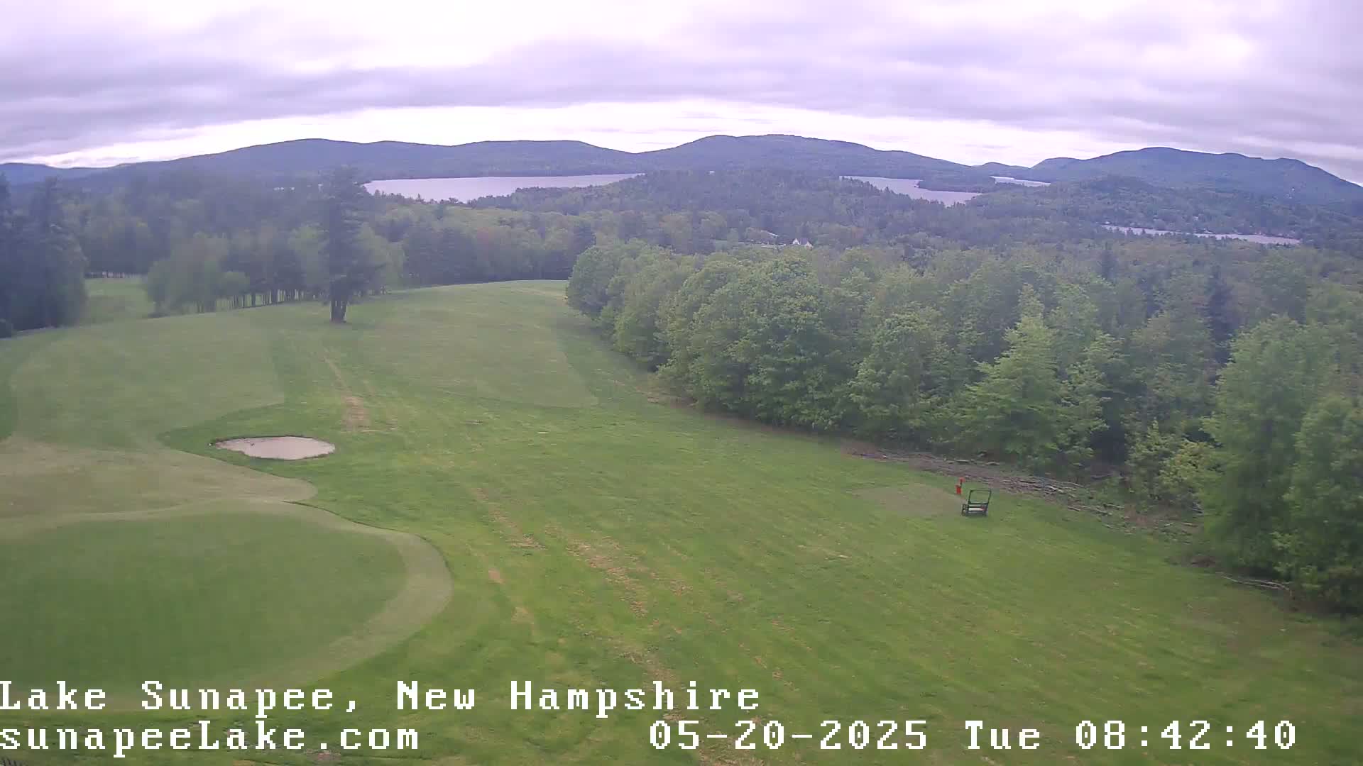An aerial view of a golf course on an overcast day shows a green fairway, a sand trap, and a distant lake and mountains.