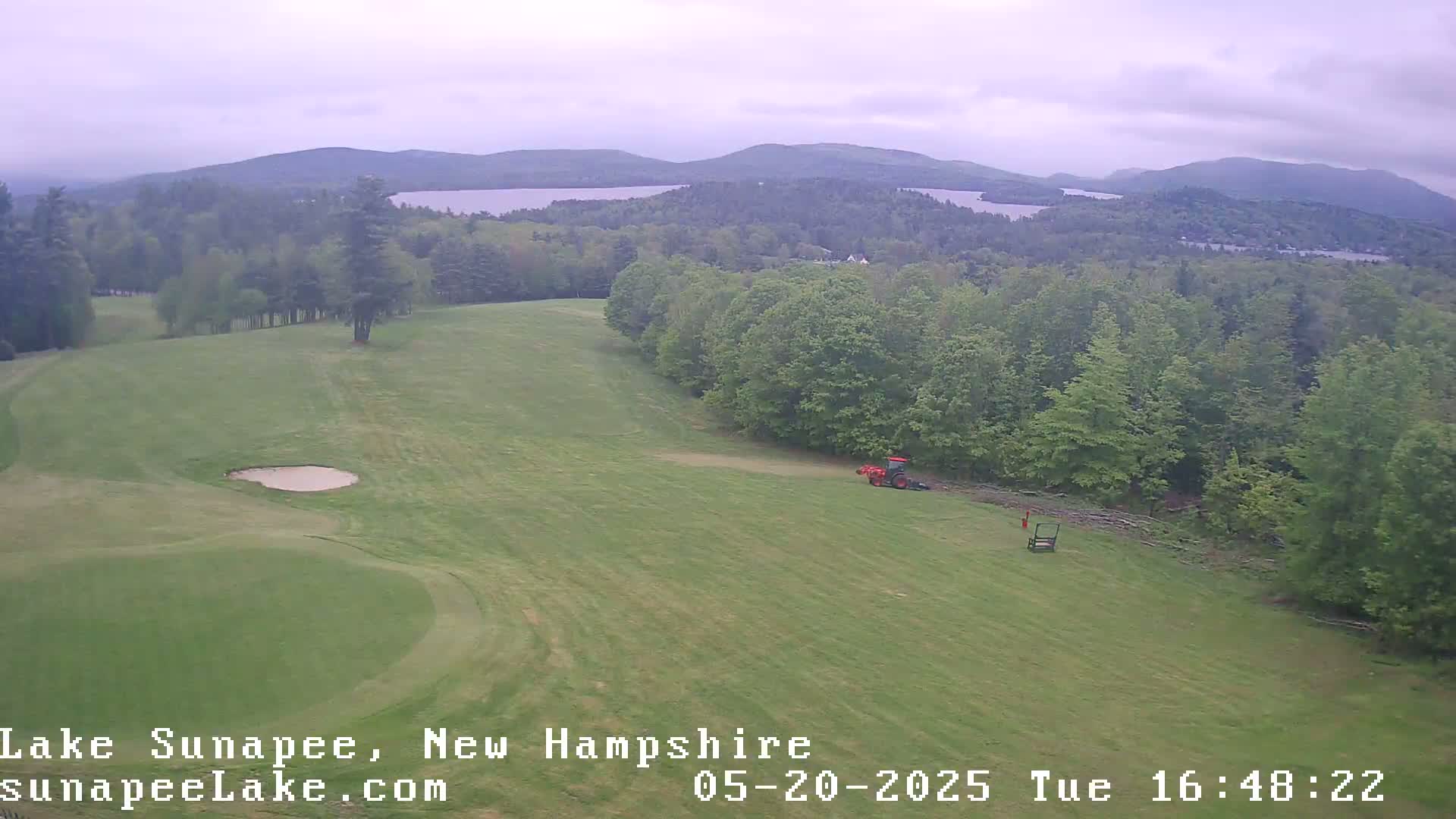 A golf course fairway slopes gently downward toward a sand trap, with a red tractor and other equipment visible in the distance, under an overcast sky and overlooking a lake and distant mountains.