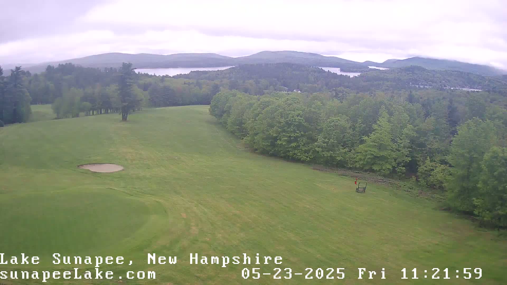 A golf course with a sand trap and distant lakes and mountains under an overcast sky.