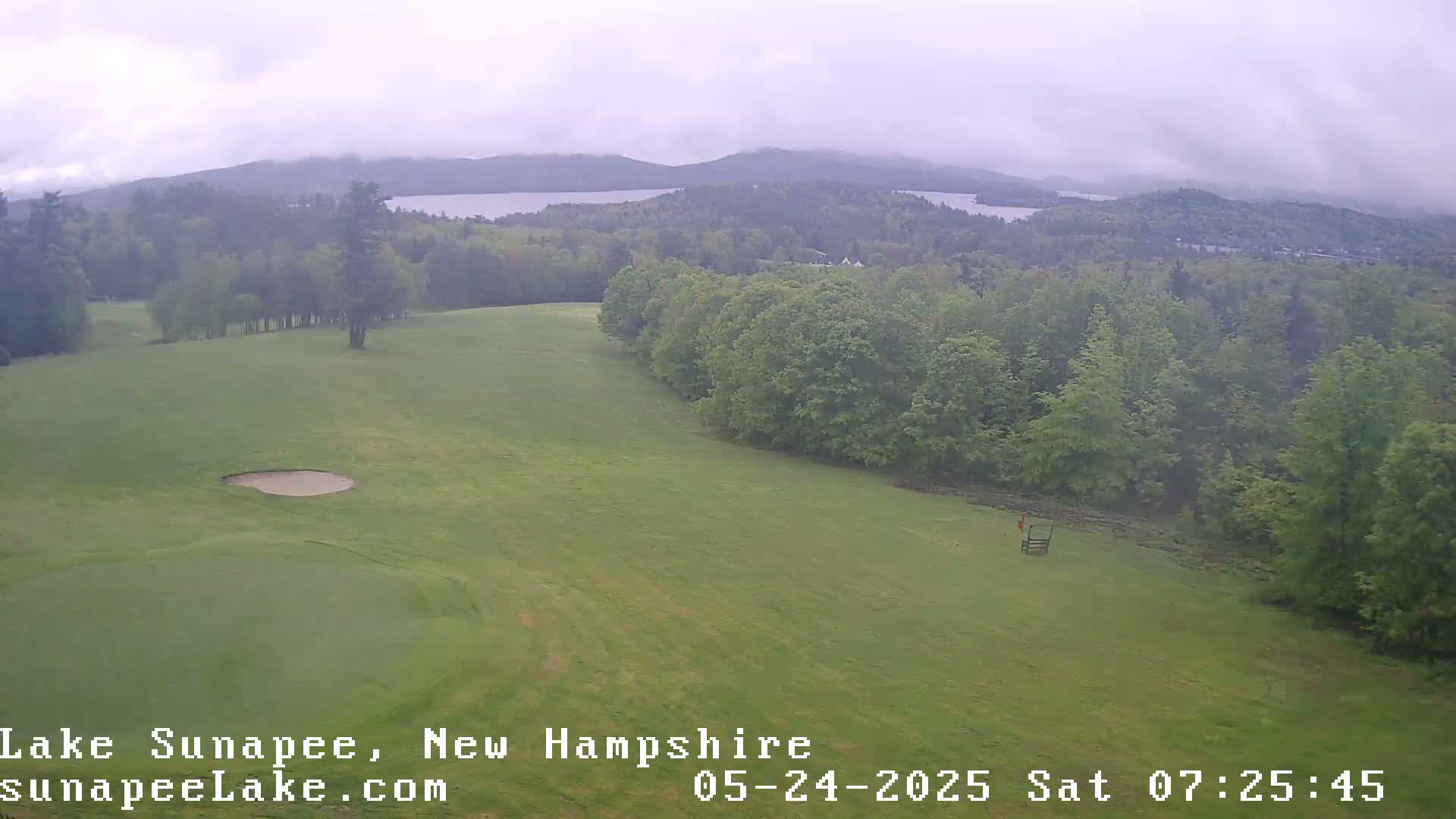 A golf course with a sand trap and a distant view of a lake and mountains under an overcast sky.