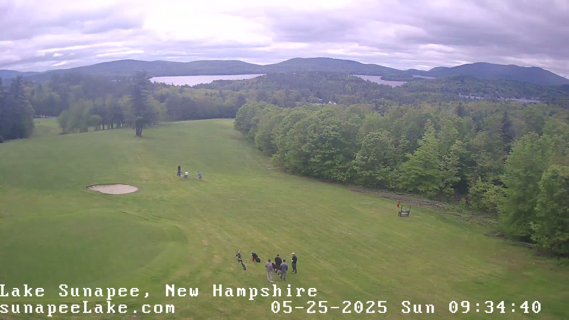 An aerial view shows a golf course with several golfers on a green fairway, overlooking a lake and forested mountains under an overcast sky.
