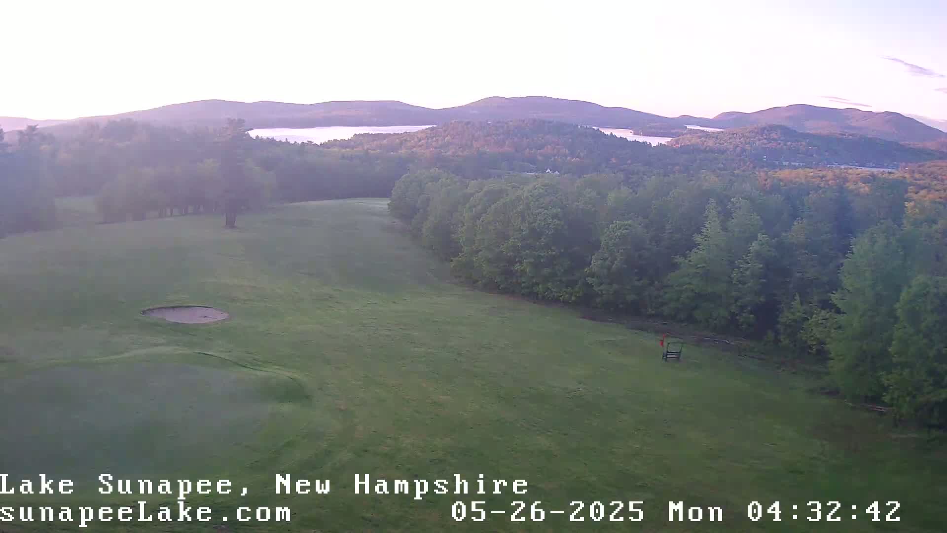 A hazy sunrise illuminates a golf course overlooking a lake and distant mountains.