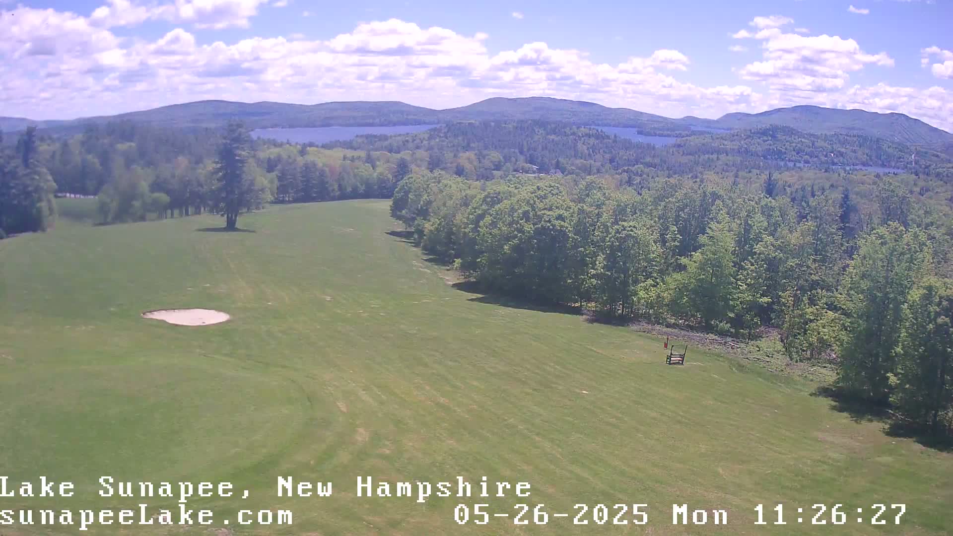 A grassy field with a sand trap, bordered by trees and overlooking a lake and distant mountains under a partly cloudy sky.