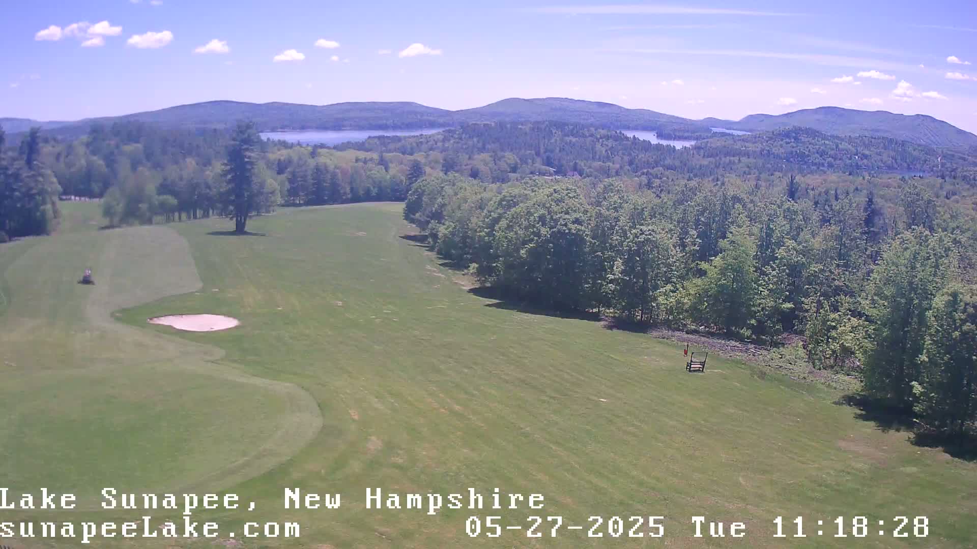 An aerial view of a golf course on a sunny day, with a lake and mountains in the distance.