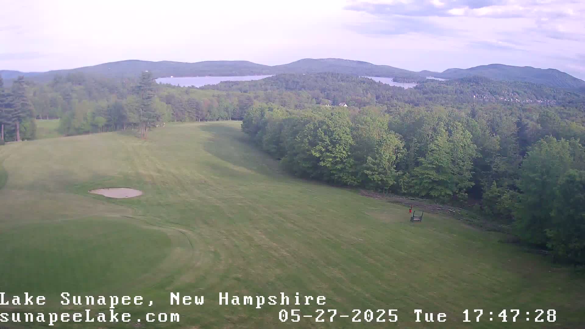 An aerial view shows a mostly-flat, grassy area of a golf course with a sand trap, a distant lake and forested mountains under a partly cloudy sky.
