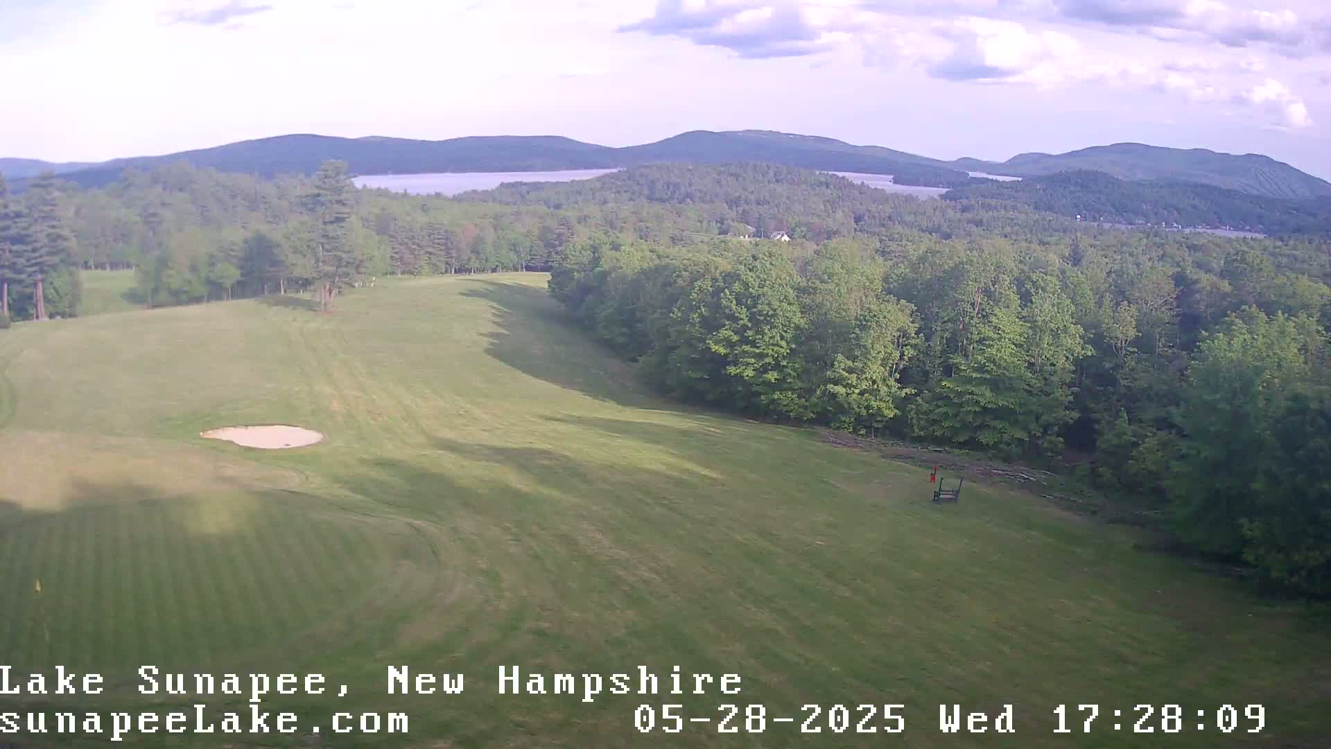 An aerial view shows a golf course with a sand trap, bordered by lush green trees and a lake and mountains in the distance under partly cloudy skies.