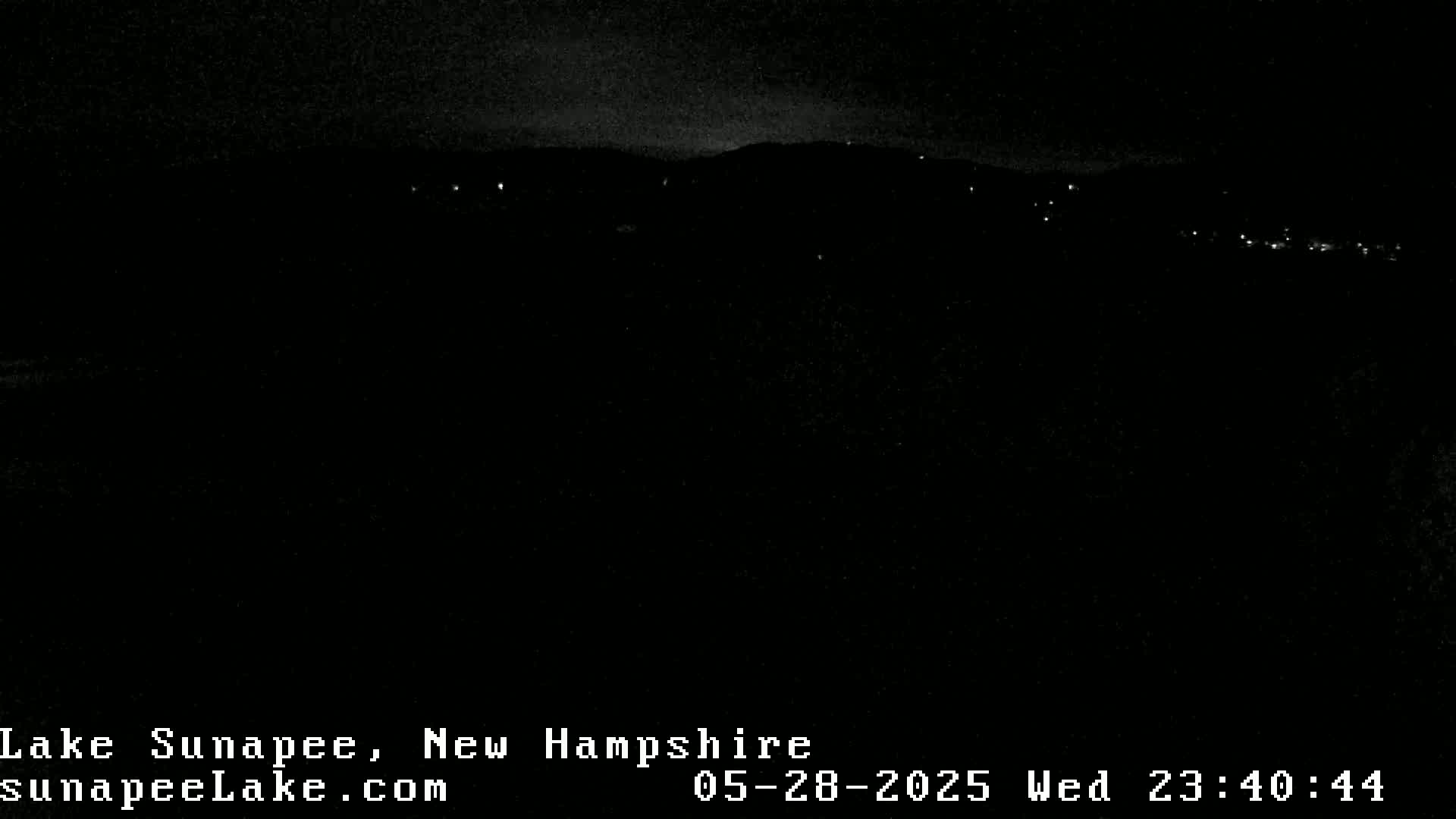 A nighttime, long shot of a dark landscape with a few distant lights visible on a hillside.