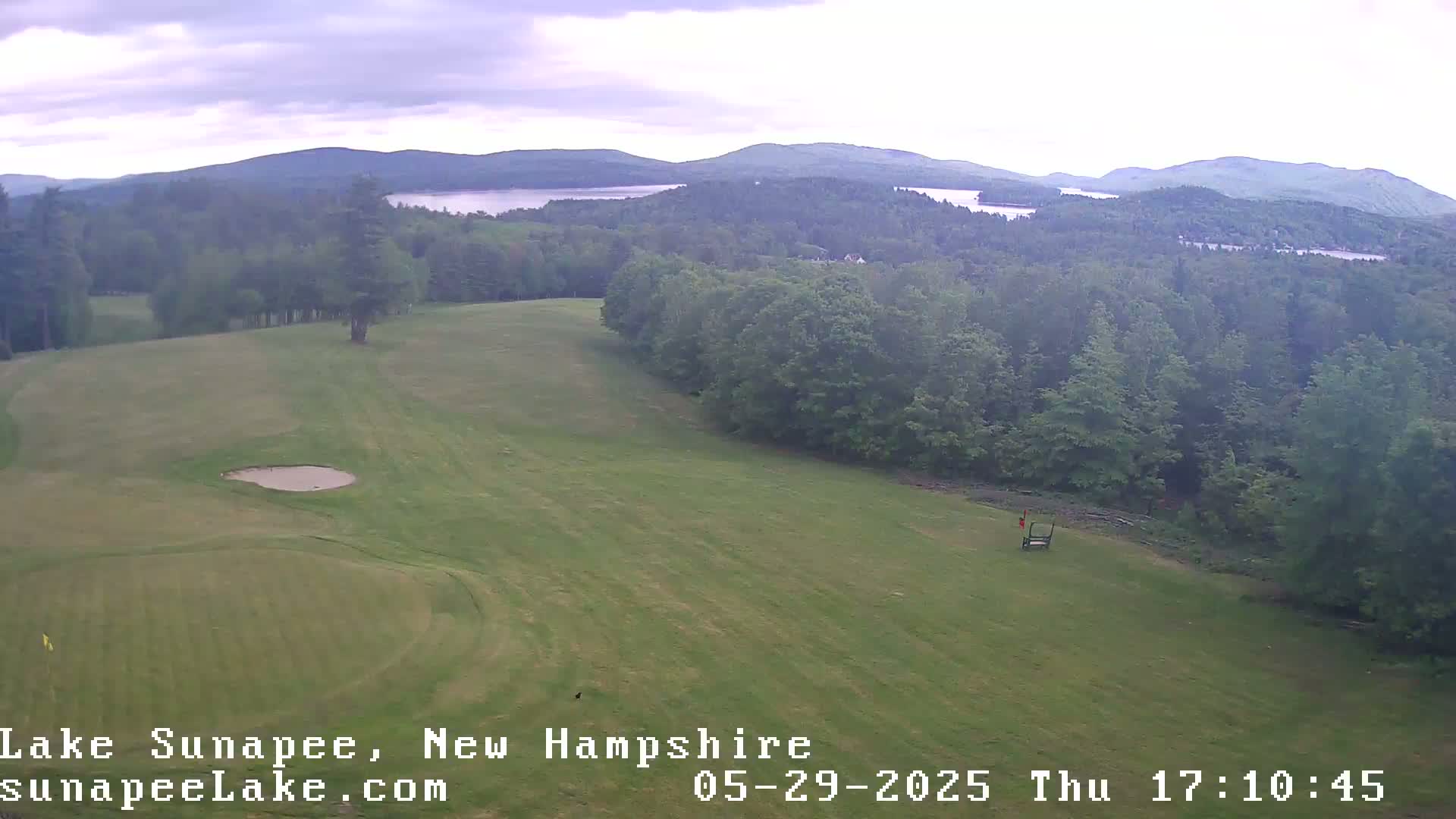 An aerial view shows a golf course with a sand trap and green, situated on a hill overlooking a wooded landscape and distant lakes under an overcast sky.