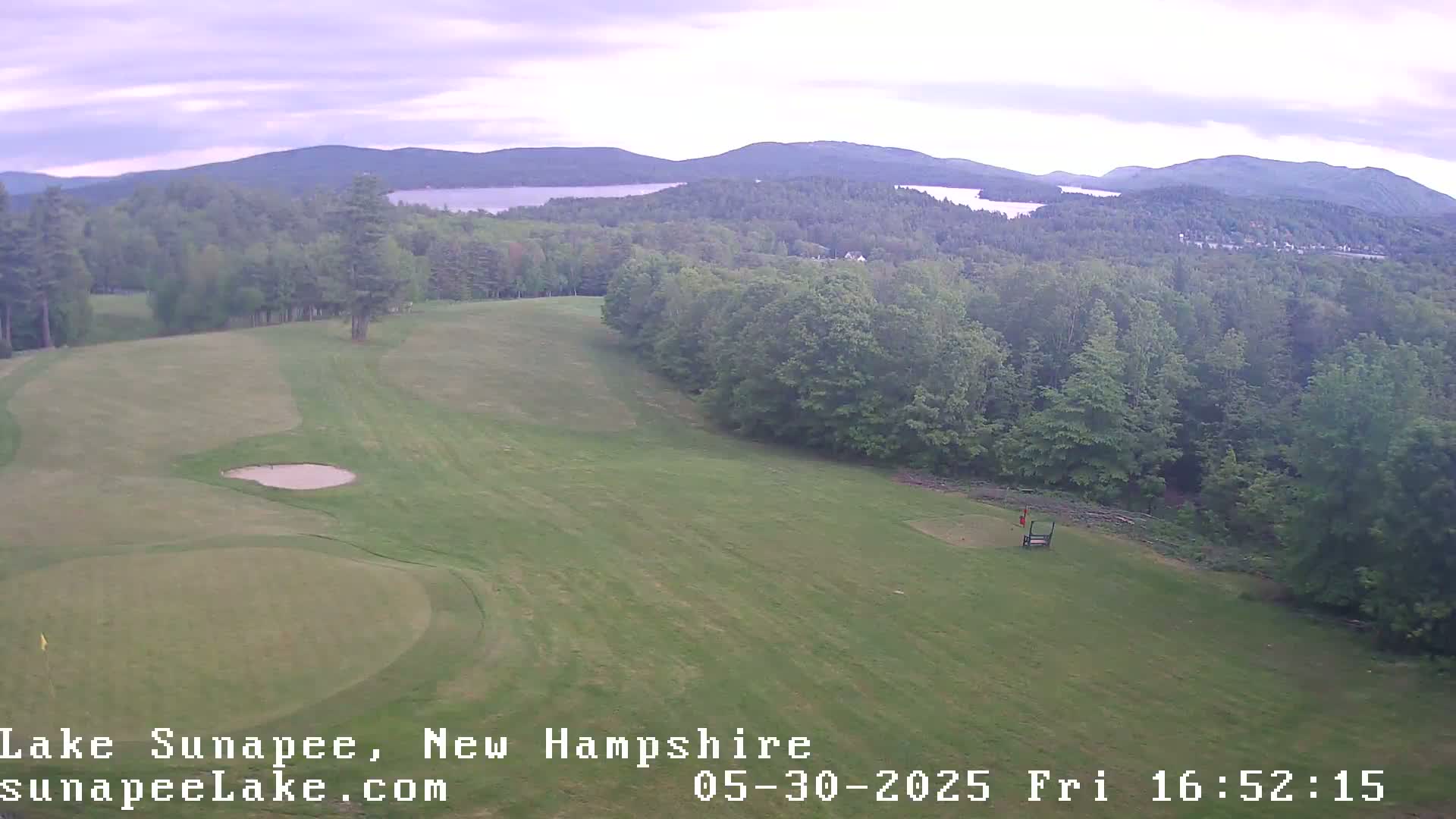 An aerial view shows a golf course with a sand trap and green, overlooking a lake and mountains under an overcast sky.