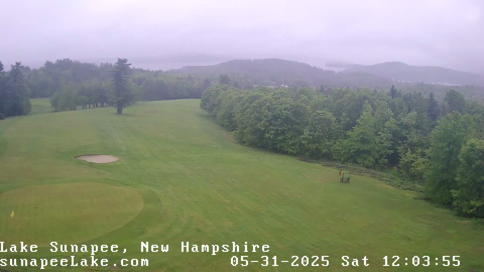 A golf course with a putting green and sand trap is shown, overlooking a misty, wooded landscape and a lake in the distance under an overcast sky.