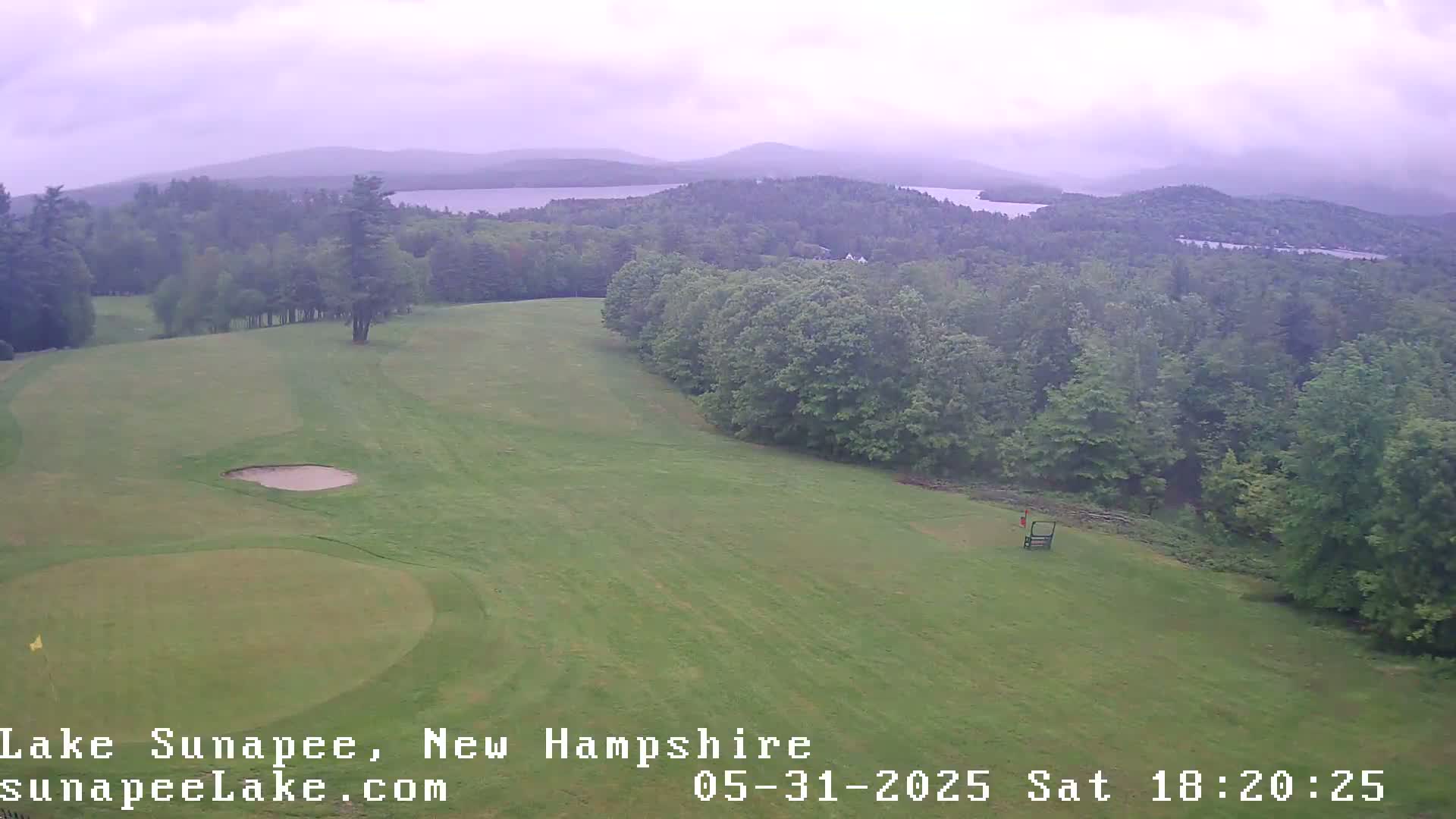 An aerial view of a golf course on a cloudy day, overlooking a lake and distant mountains.