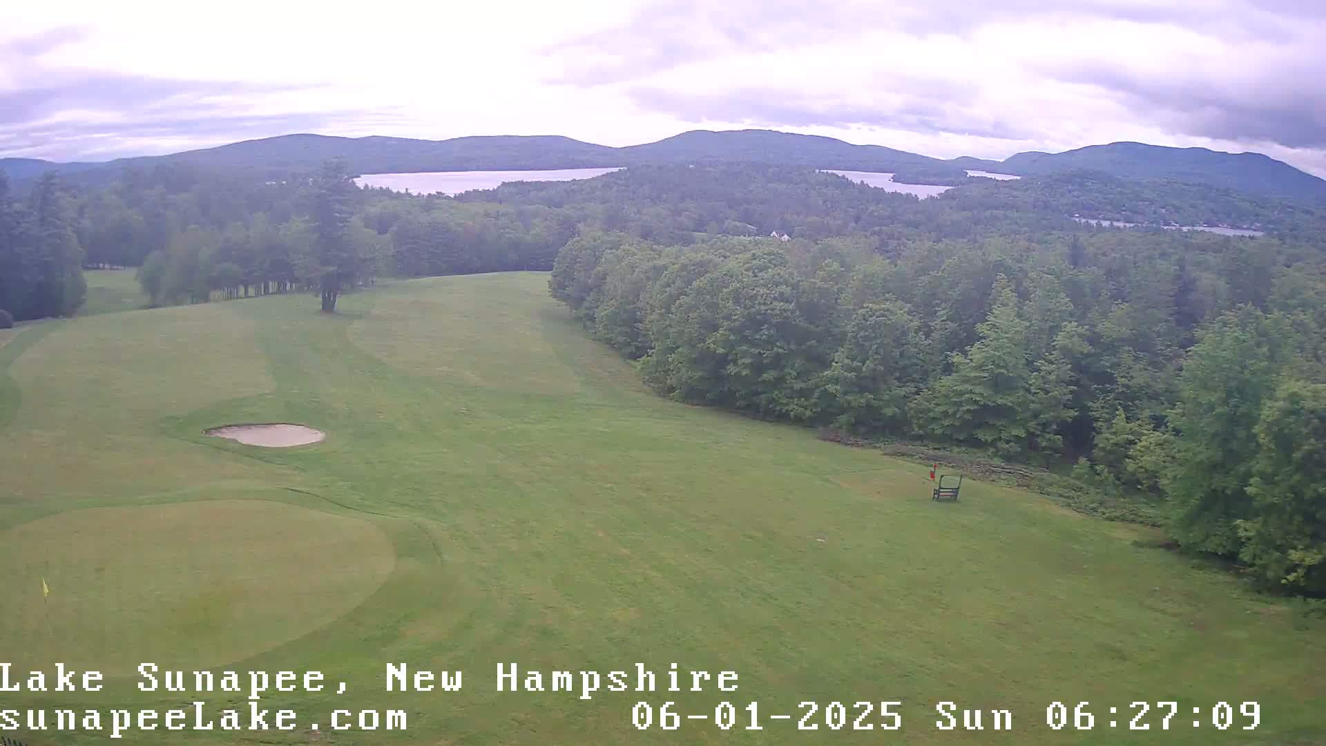 An aerial view shows a golf course with a green and sand trap, bordered by a line of trees, overlooking a lake and distant mountains under an overcast sky.