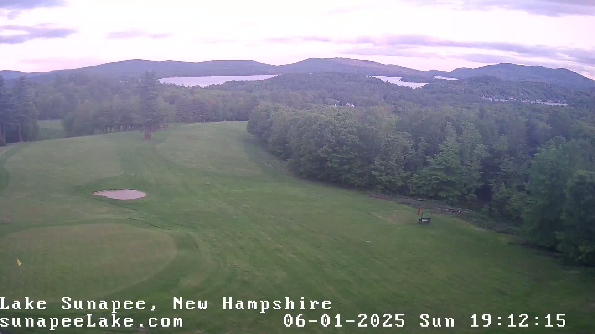 An elevated view shows a golf course with a small pond, surrounded by lush green trees and distant mountains under a partly cloudy sky.