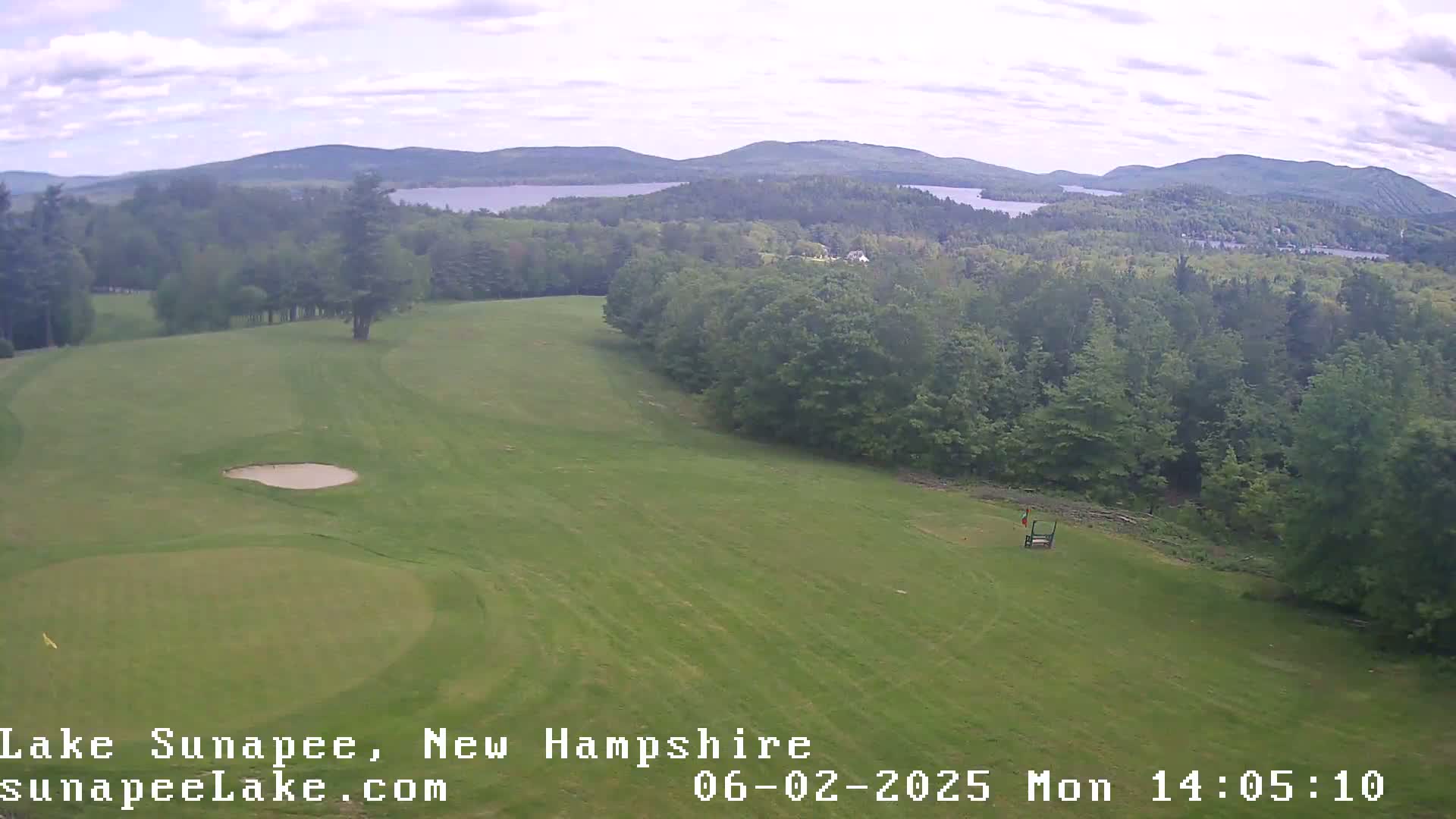 An elevated view shows a golf course with a sand trap and green, overlooking a wooded area and a lake under a partly cloudy sky.