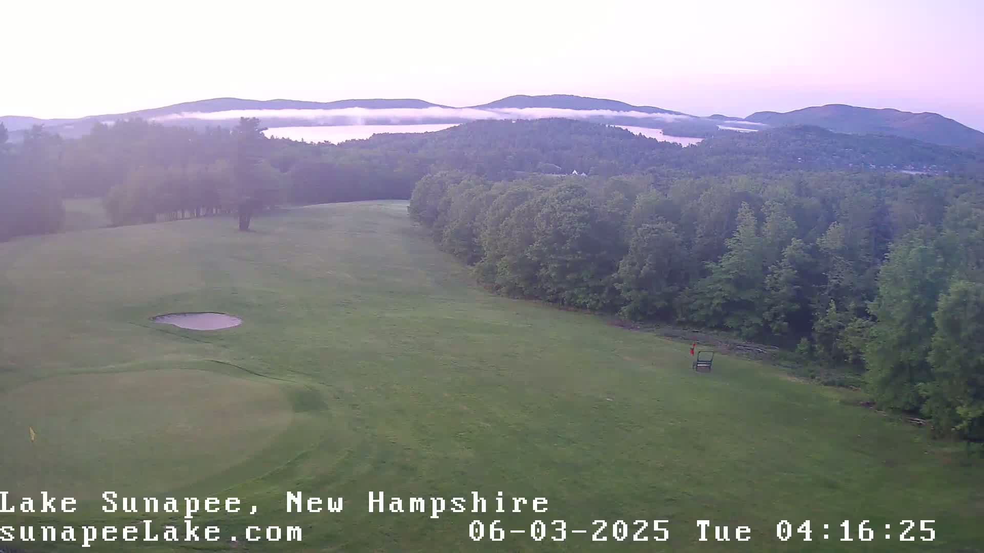 A golf course with a small body of water, set against a background of misty, forested hills under a hazy sky.