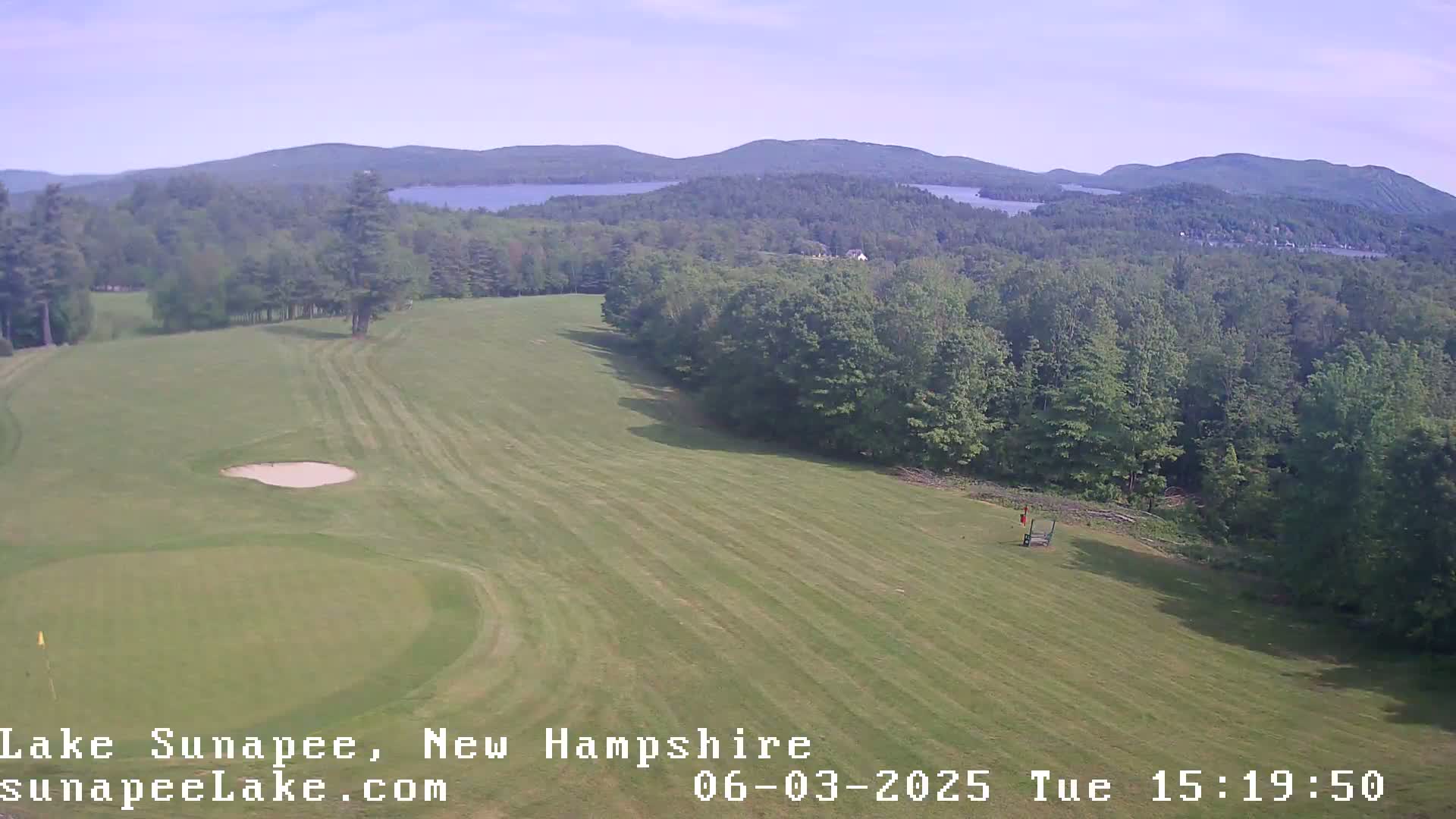 An aerial view shows a golf course with a sand trap and green, bordered by a wooded area and distant lakes under a clear sky.