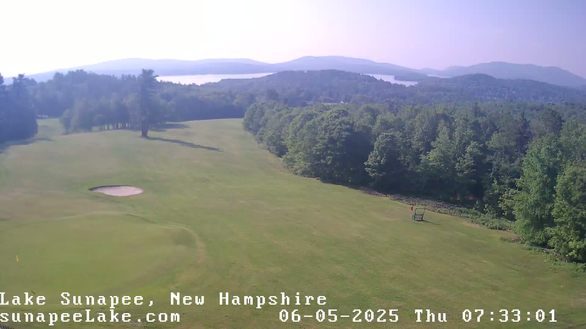 A golf course with a sand trap and distant lake and mountains is visible under hazy, sunny conditions.