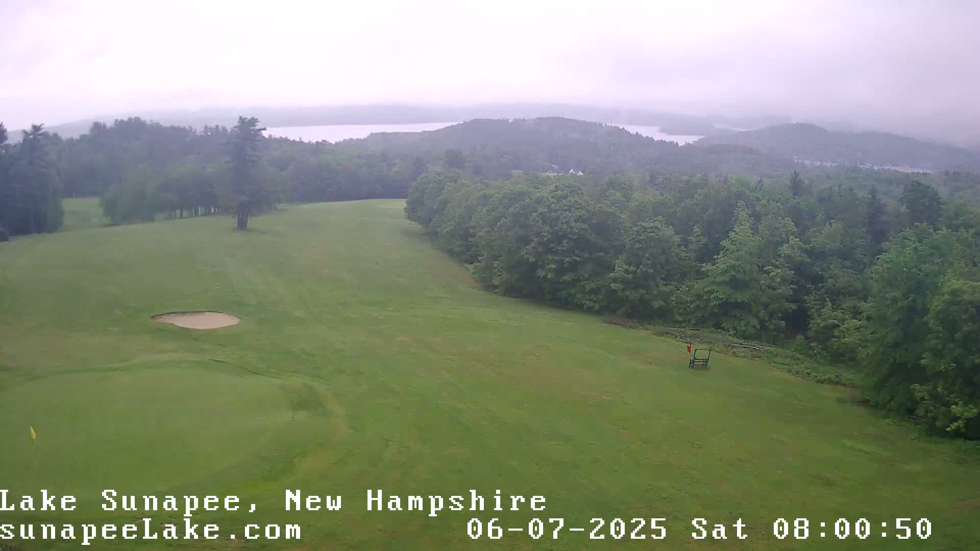 A golf course with a sand trap, overlooking a hazy, wooded landscape and a distant lake under overcast skies.
