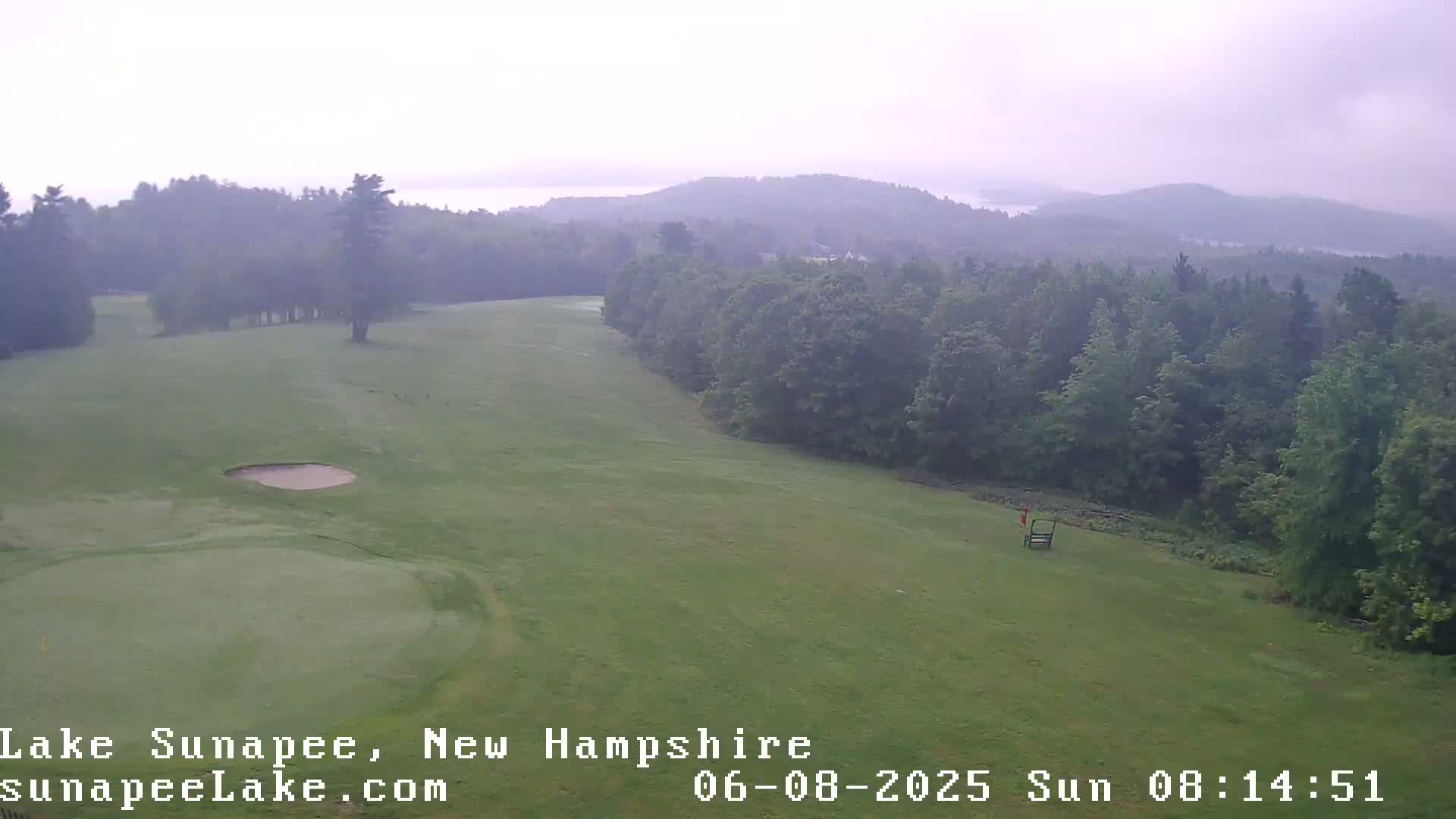 A golf course with a sand trap and flag is visible, surrounded by a hazy, forested landscape under an overcast sky.