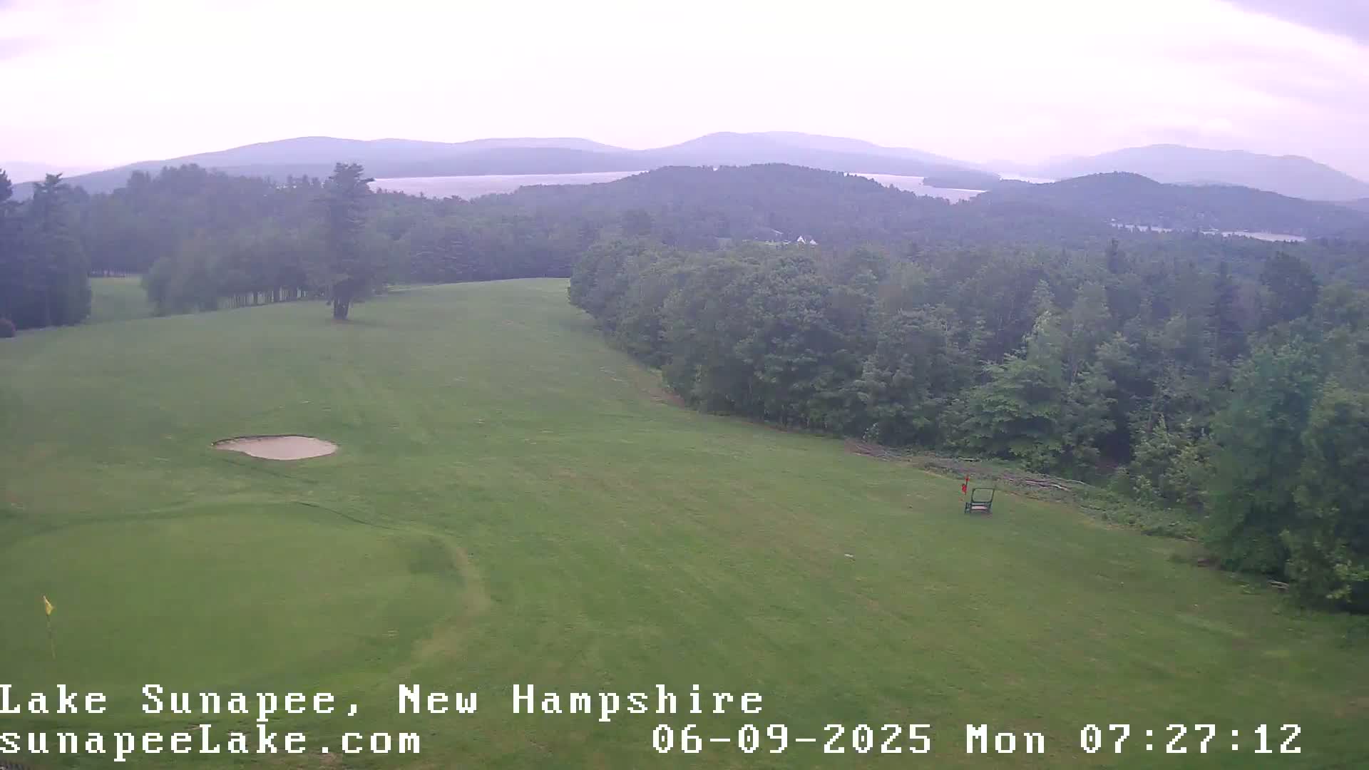 An elevated view shows a golf course with a sand trap and a golf cart, overlooking a hazy, mountainous landscape with a lake in the distance.