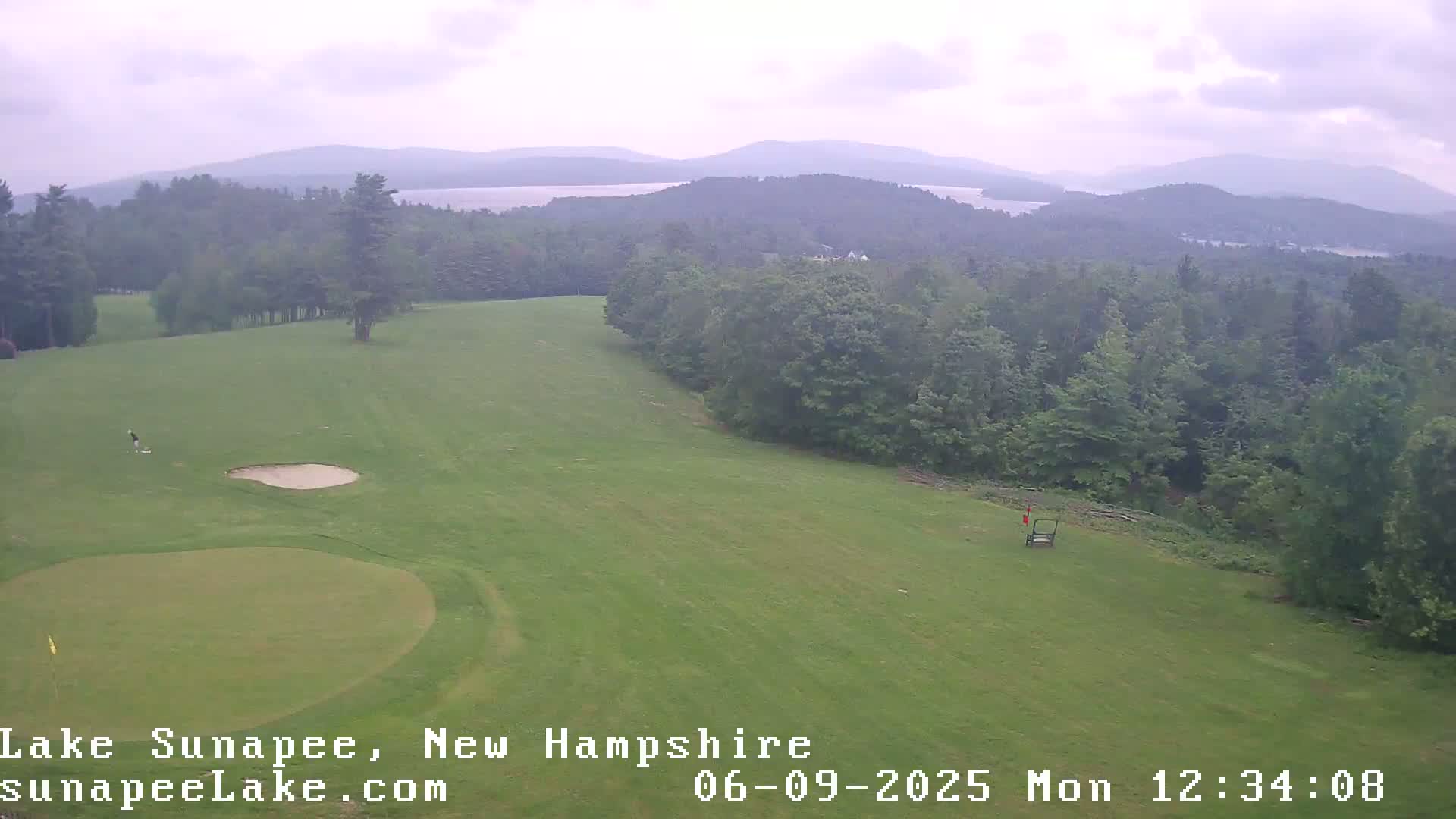 A golfer is seen on a green golf course overlooking a lake and distant mountains under an overcast sky.