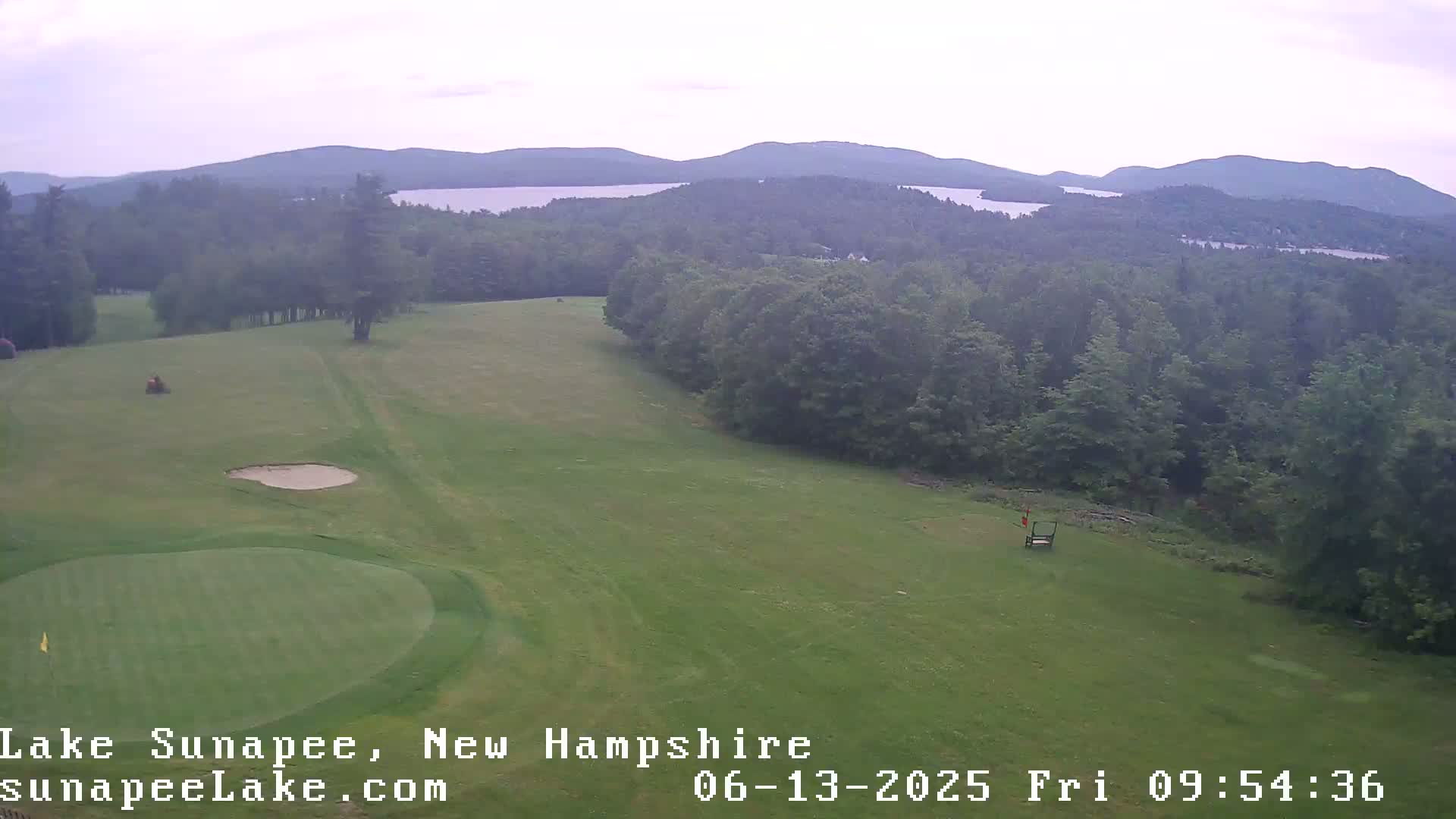 An aerial view shows a golf course green with a sand trap and distant mountains under an overcast sky.