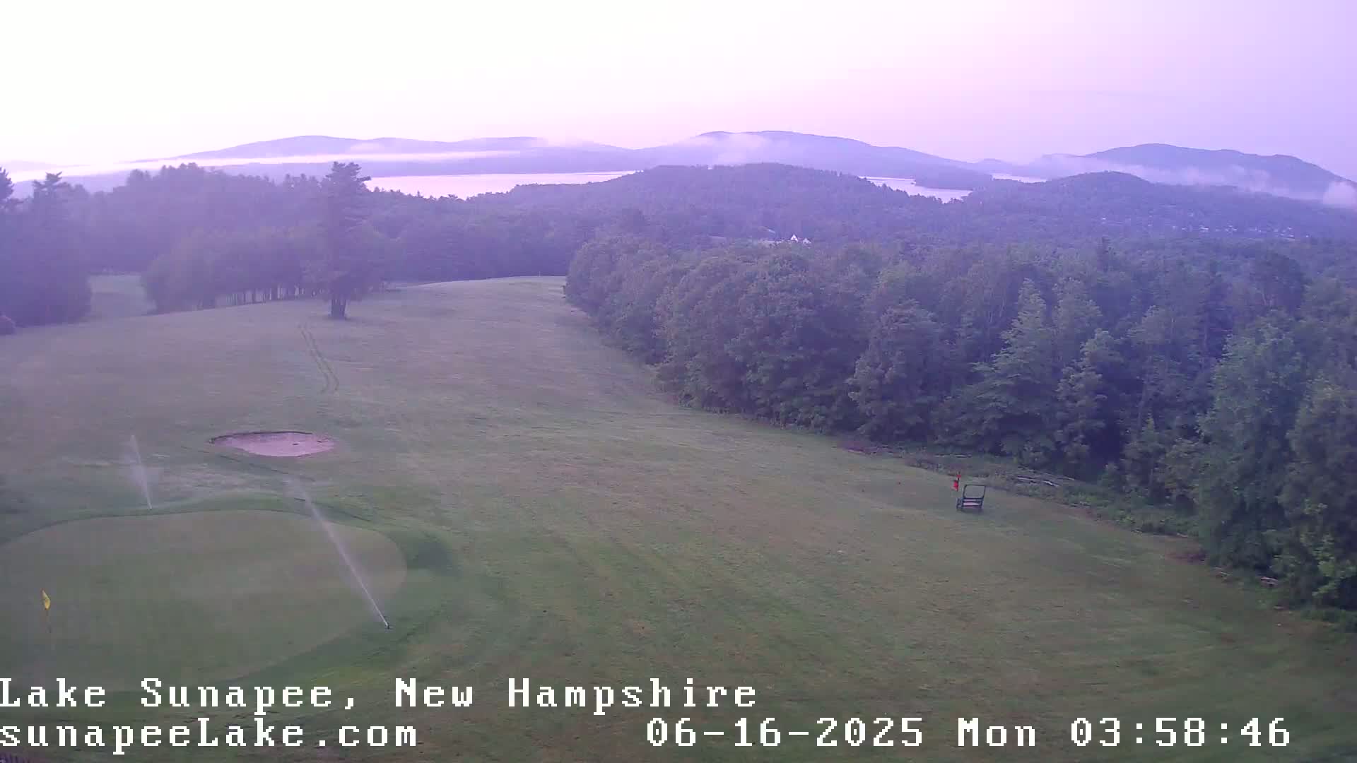 An aerial view shows a golf course with sprinklers active, overlooking a misty lake and forested hills under a pale purple sky.
