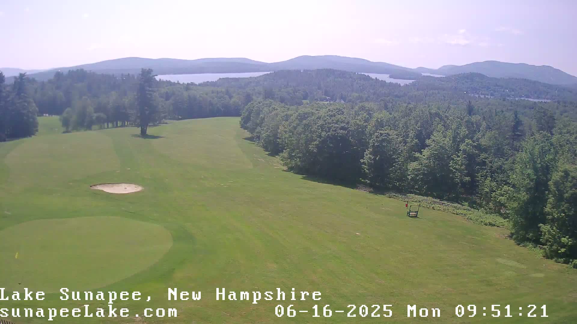 An aerial view shows a green golf course on a sunny day with a lake and distant mountains visible in the background.