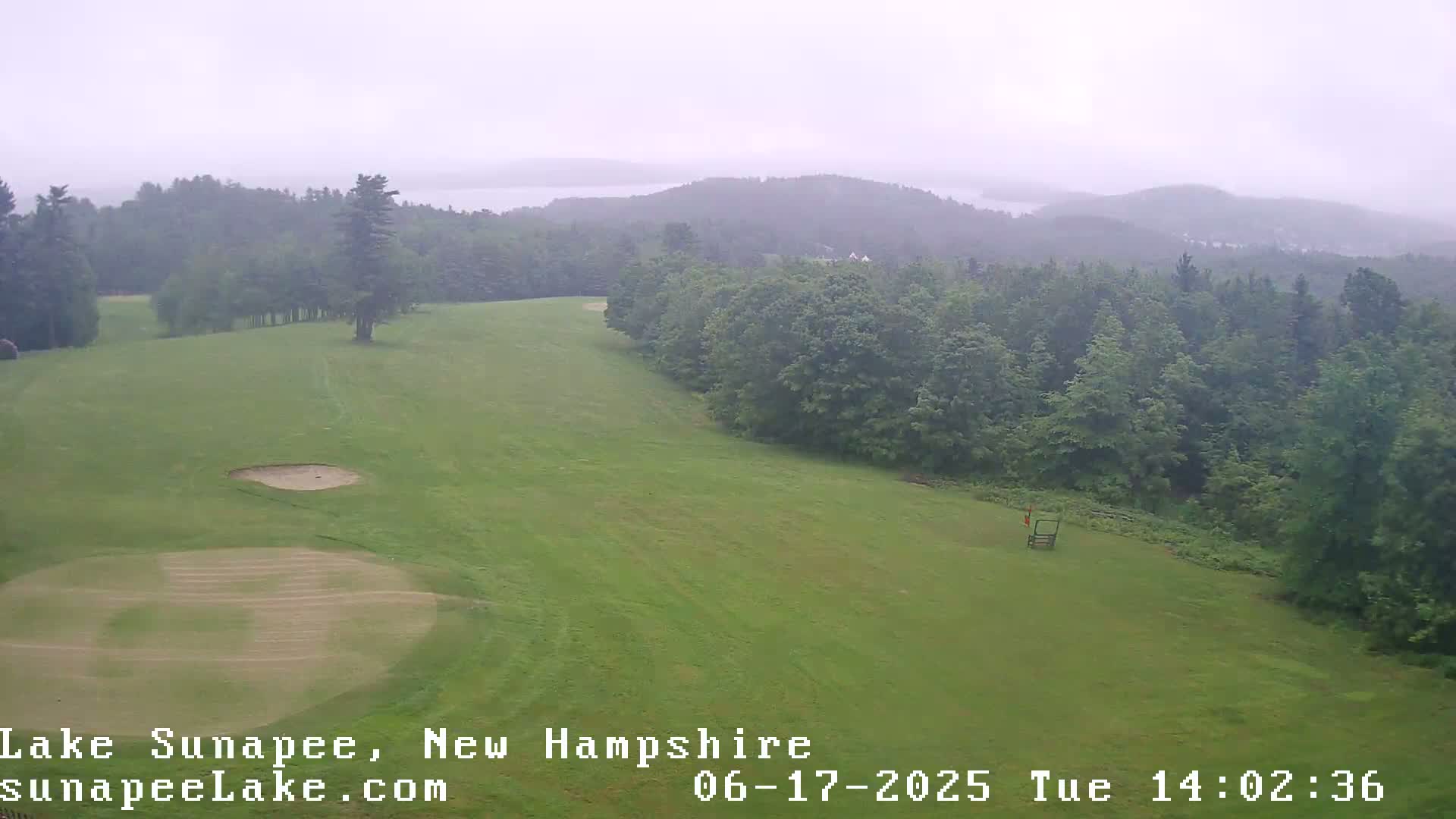 A golf course with sand traps and a hazy view of a lake and distant hills under an overcast sky.