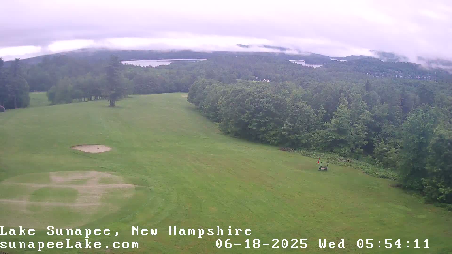 A golf course with sand traps and a flag is visible in the foreground, overlooking a lake and forested hills shrouded in a misty, overcast sky.