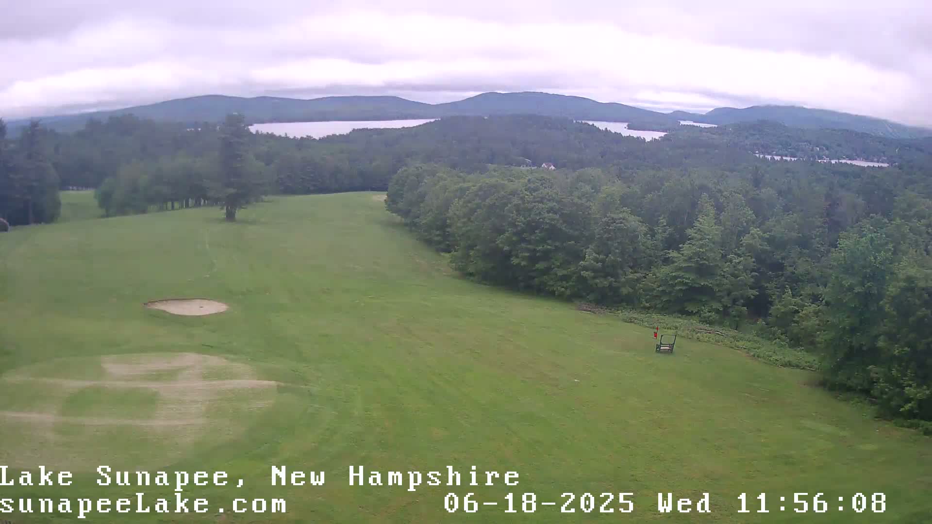 An aerial view shows a golf course with a sand trap, overlooking a lake and mountains under an overcast sky.