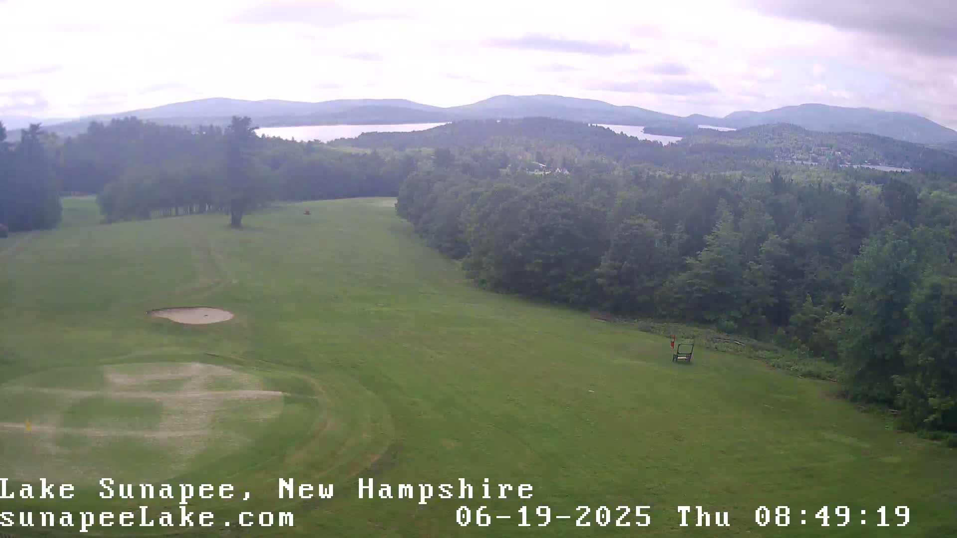 A mostly cloudy day overlooks a golf course, with a distant lake and mountains visible in the background.