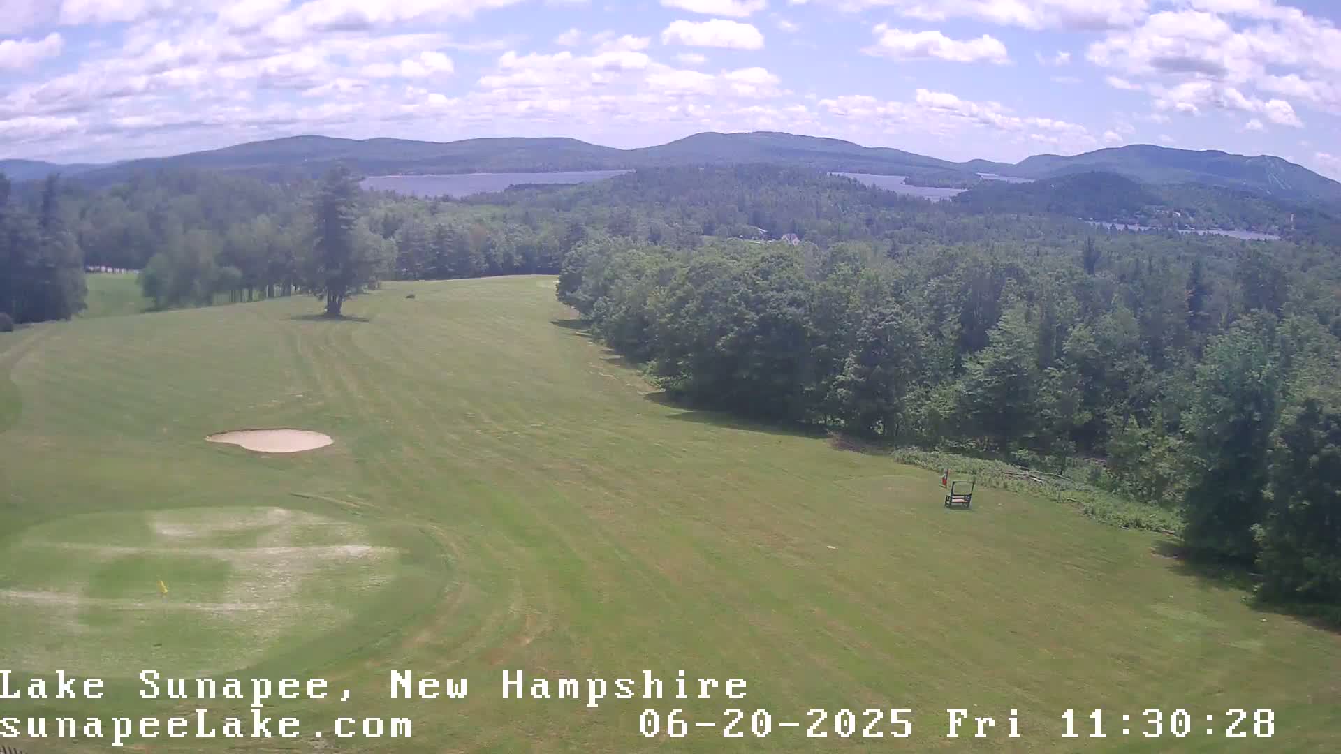 A golf course fairway stretches towards a lake and distant mountains under a partly cloudy sky.