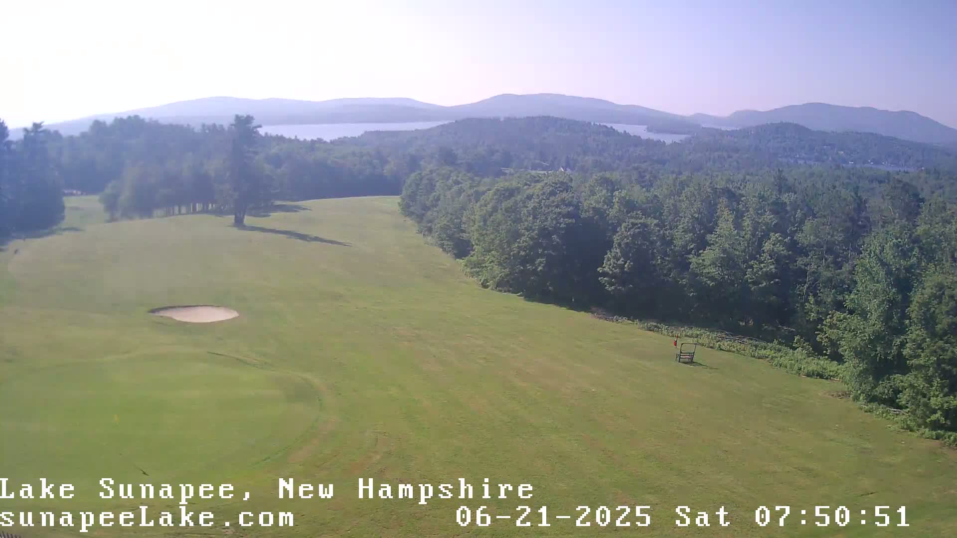 A high-angle view shows a golf course on a sunny day, with a lake and mountains visible in the distance.