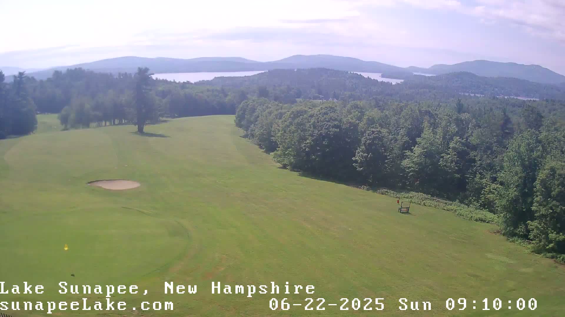 An aerial view shows a golf course on a sunny day, with a lake and mountains visible in the distance.