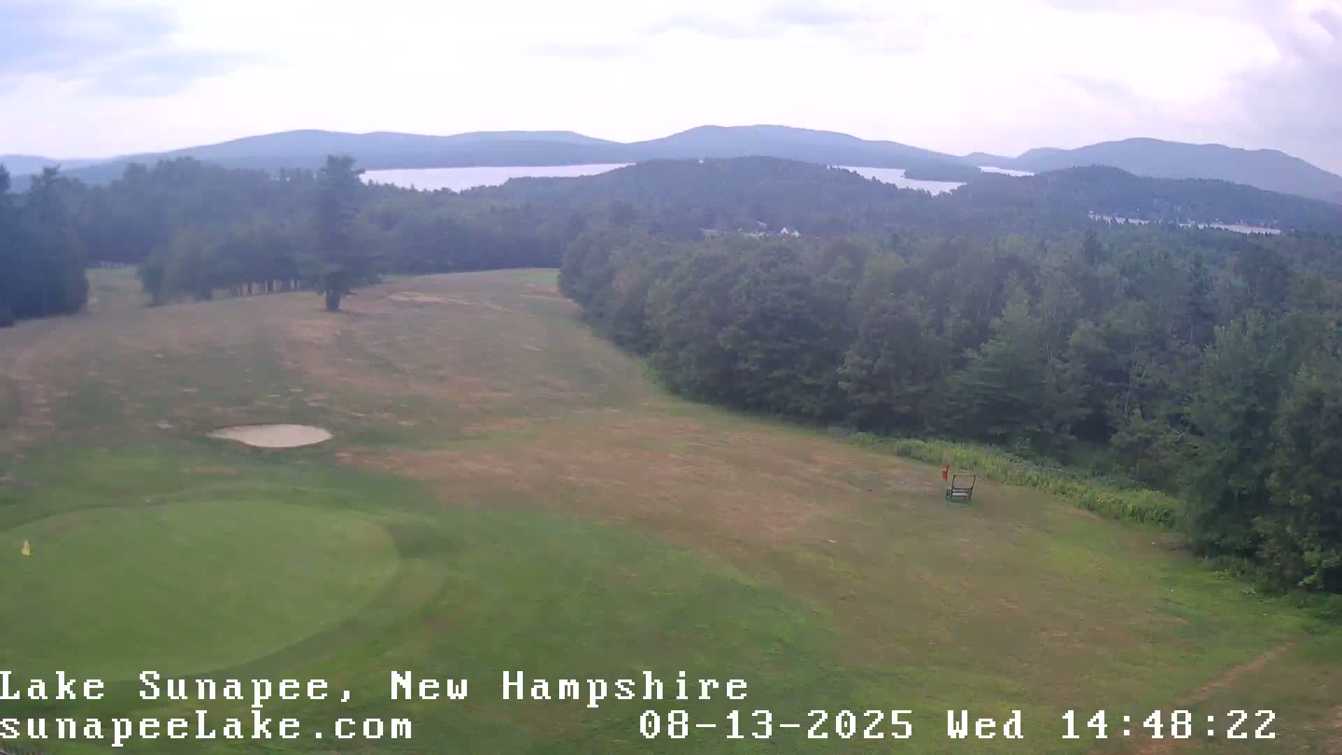An elevated view shows a golf course with a sand trap and green, bordered by trees and overlooking a hazy lake and distant mountains under a cloudy sky.