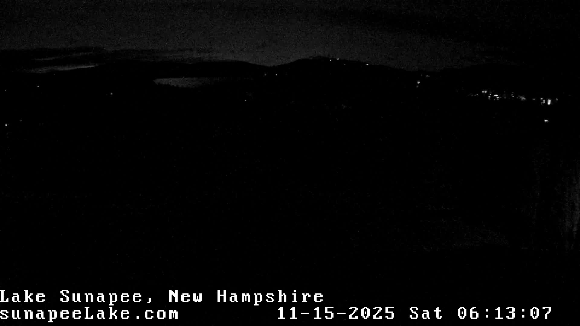 The image displays an extremely dark outdoor night scene, revealing only faint silhouettes of distant land or hills dotted with scattered lights under a dark, possibly overcast sky.