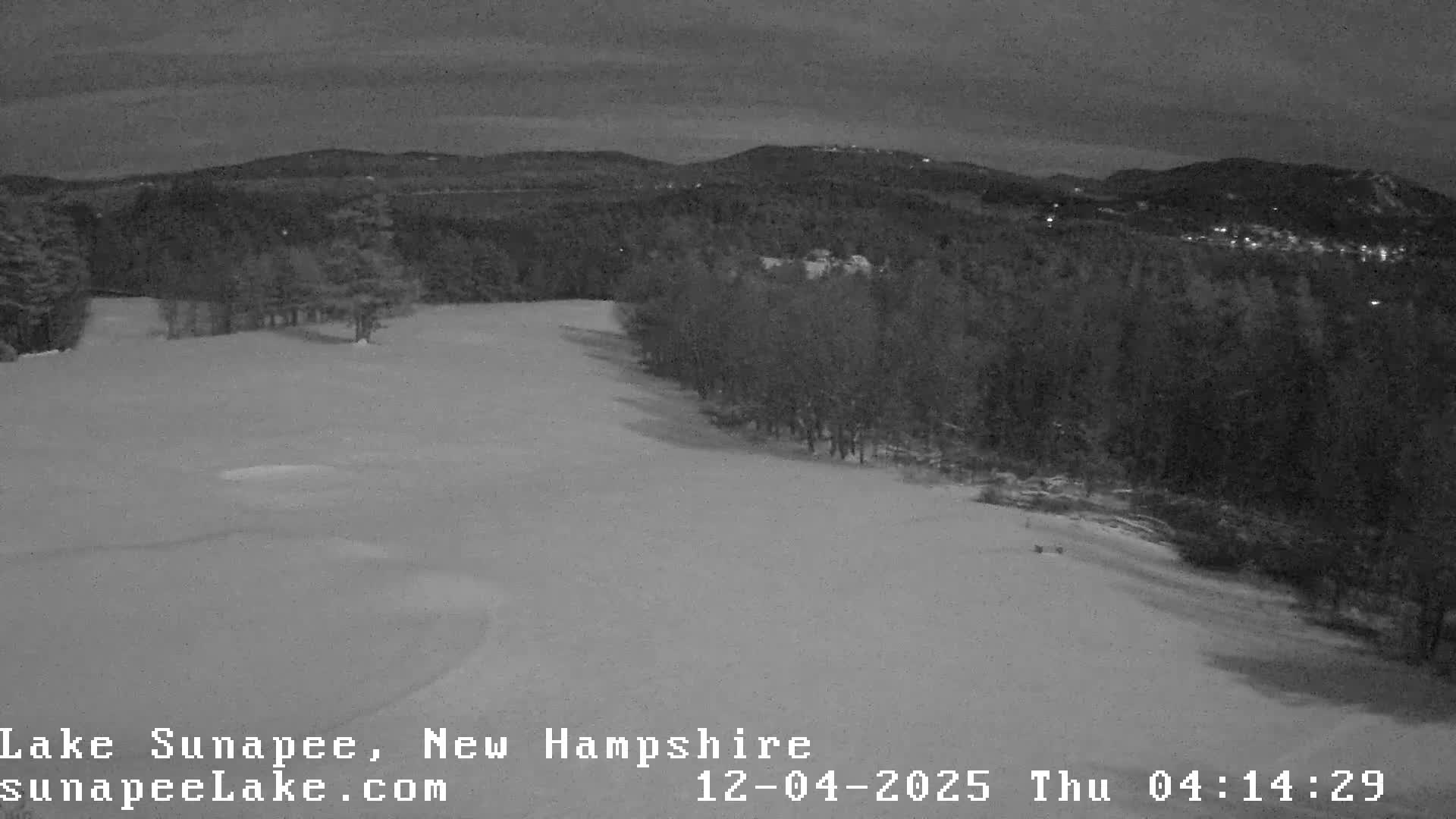 A wide, snow-covered landscape at night features a clearing in the foreground, bordered by a dense treeline, with distant, forested hills showing scattered lights under a dark, clear sky.