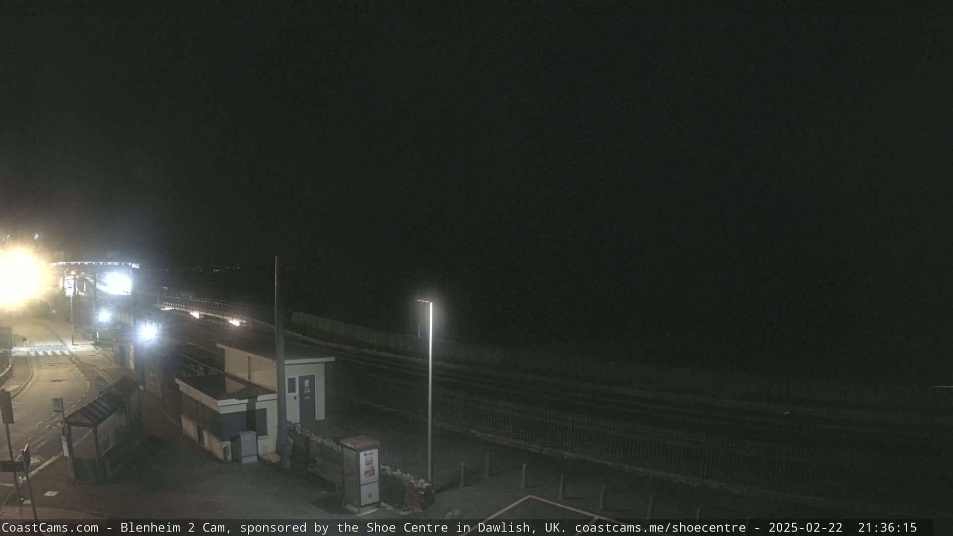 A nighttime view of a mostly empty roadway and train tracks next to a small building, under a dark, clear sky.