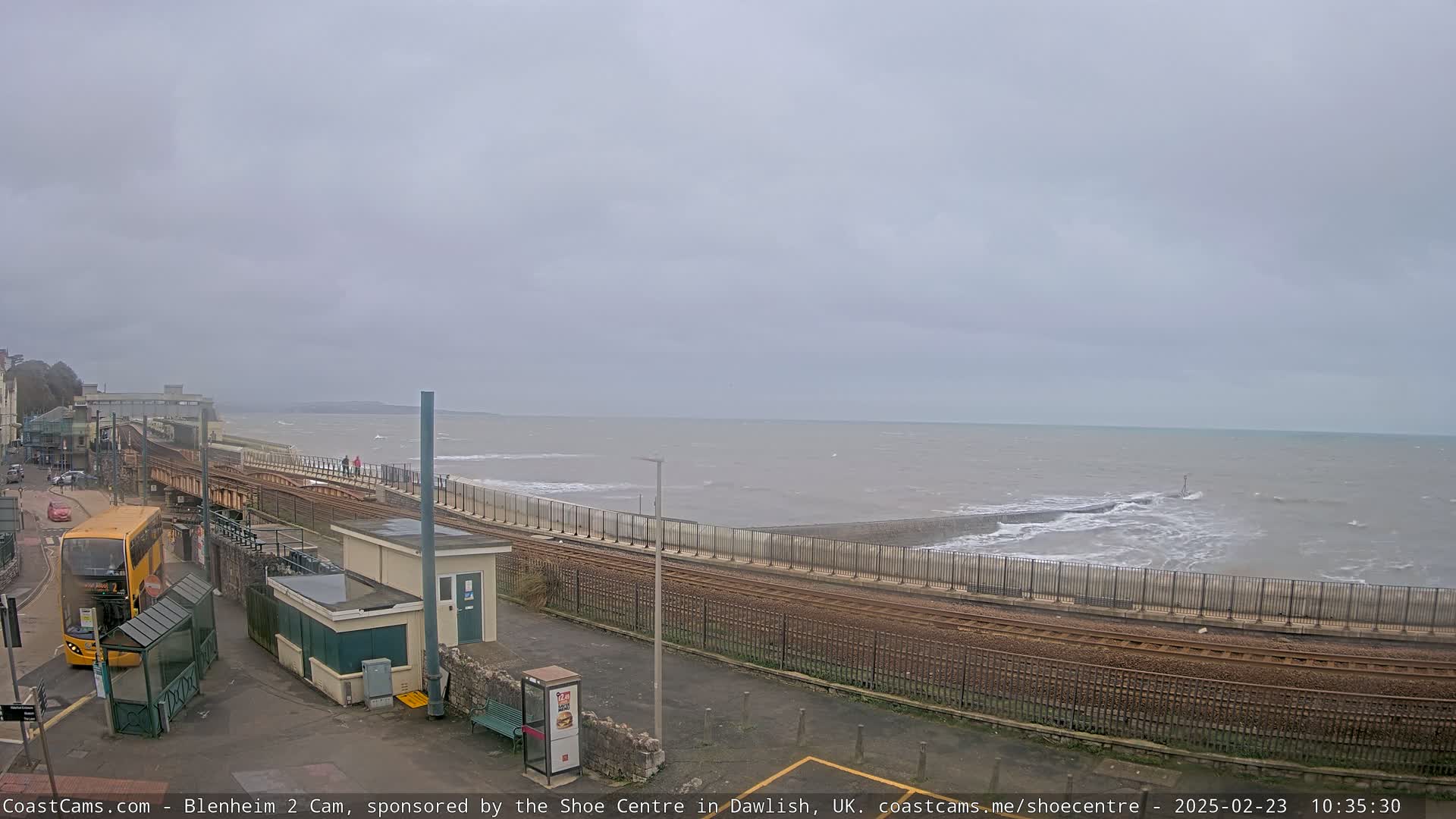 A train track runs alongside a seawall separating a bus stop and buildings from a choppy sea under an overcast sky.